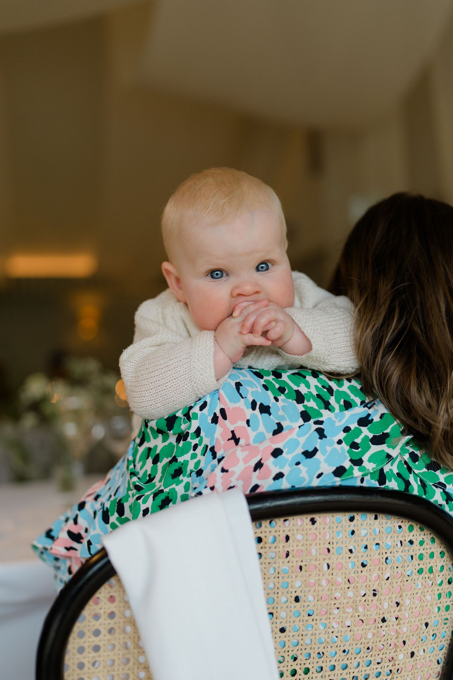 Adorable blue-eyed baby in cream sweater rests on mothers shoulder at a wedding reception.