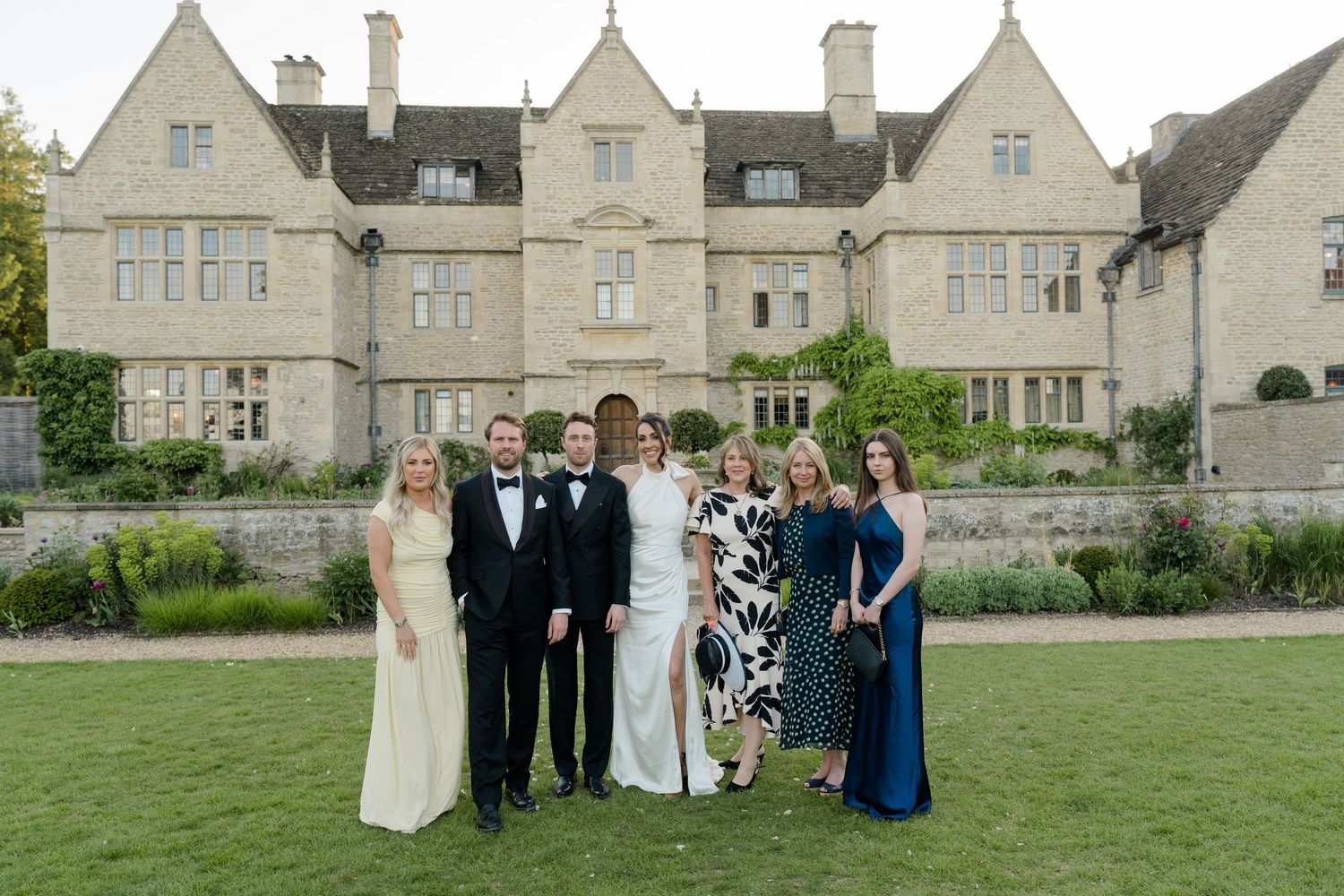 Wedding party poses in formal attire in front of a grand stone manor house at dusk.