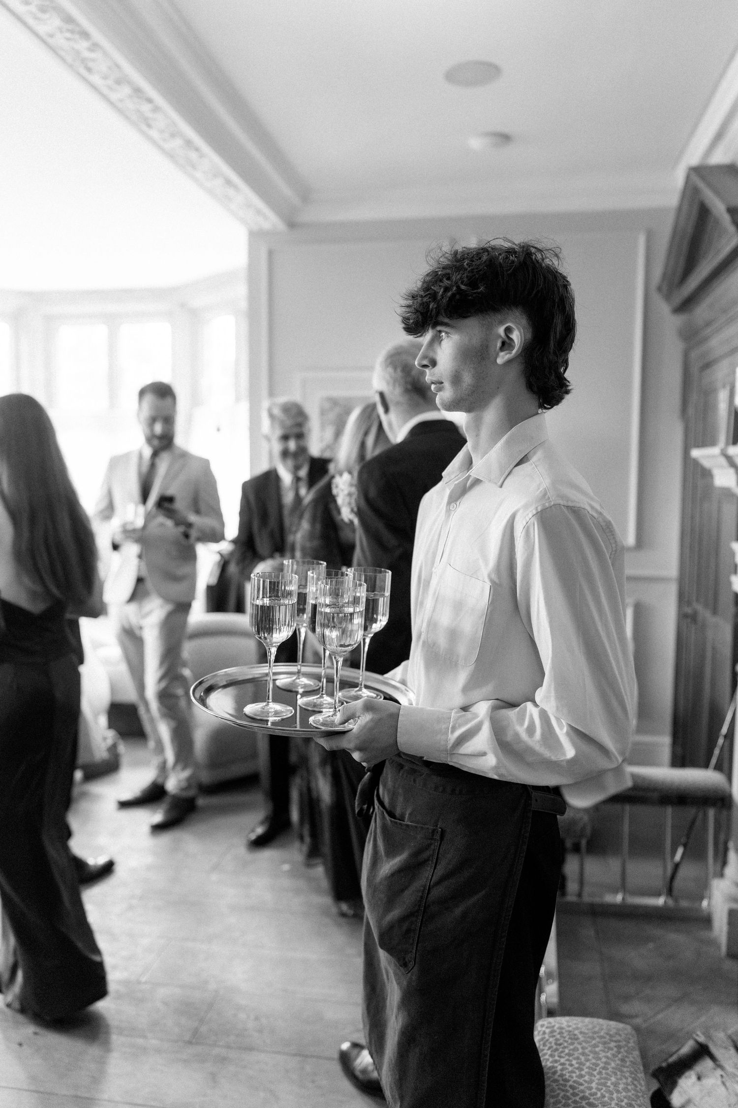 Black and white photo of young waiter holding champagne glasses on silver tray at elegant wedding reception.