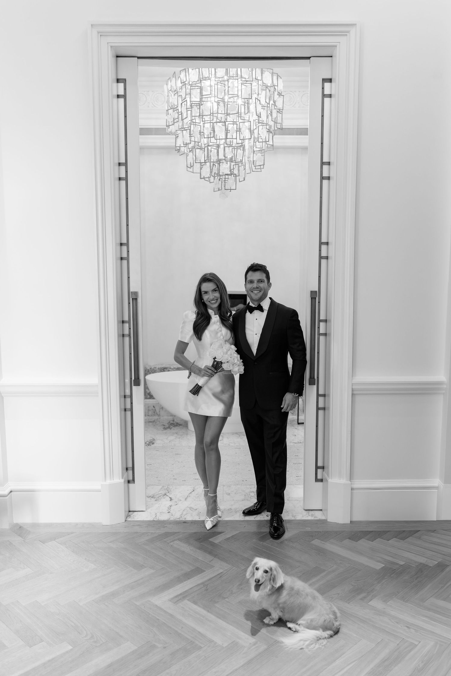 Elegant couple in wedding attire posing in doorway with chandelier and golden retriever dog in black and white photo.
