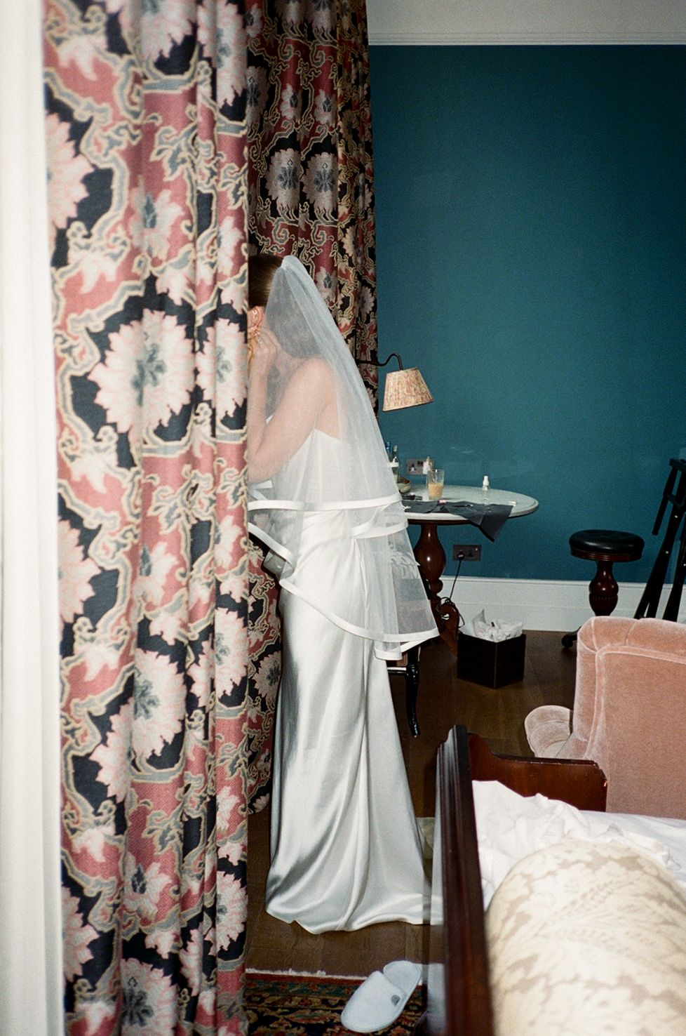 Bride in white satin gown and veil stands by ornate floral curtains in teal-walled room preparing for wedding.