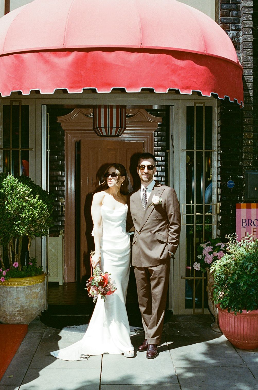 Bride and groom posing under a red awning, both wearing sunglasses, bride holding a colorful bouquet.