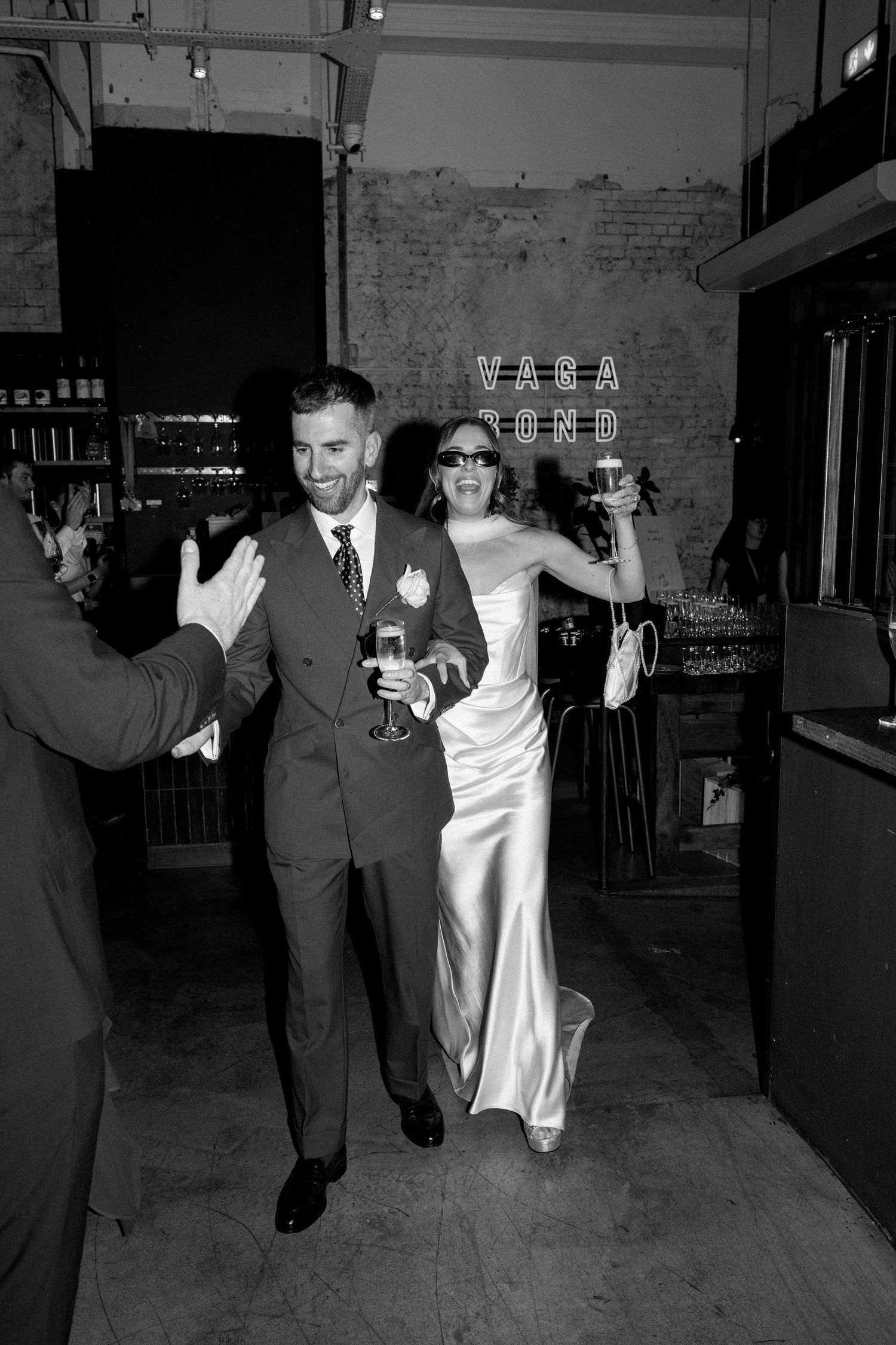 Black and white photo of smiling bride and groom entering wedding reception venue holding champagne glasses.