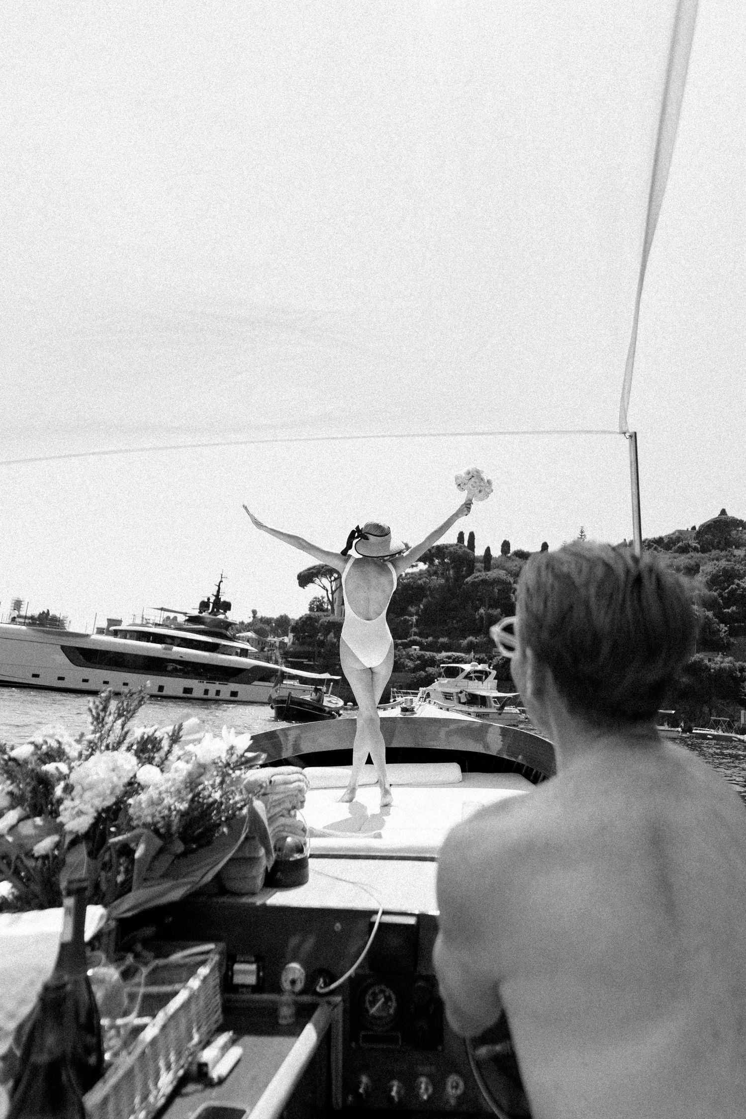 Joyful bride in white swimsuit stands on yacht bow with arms raised while groom watches from deck.