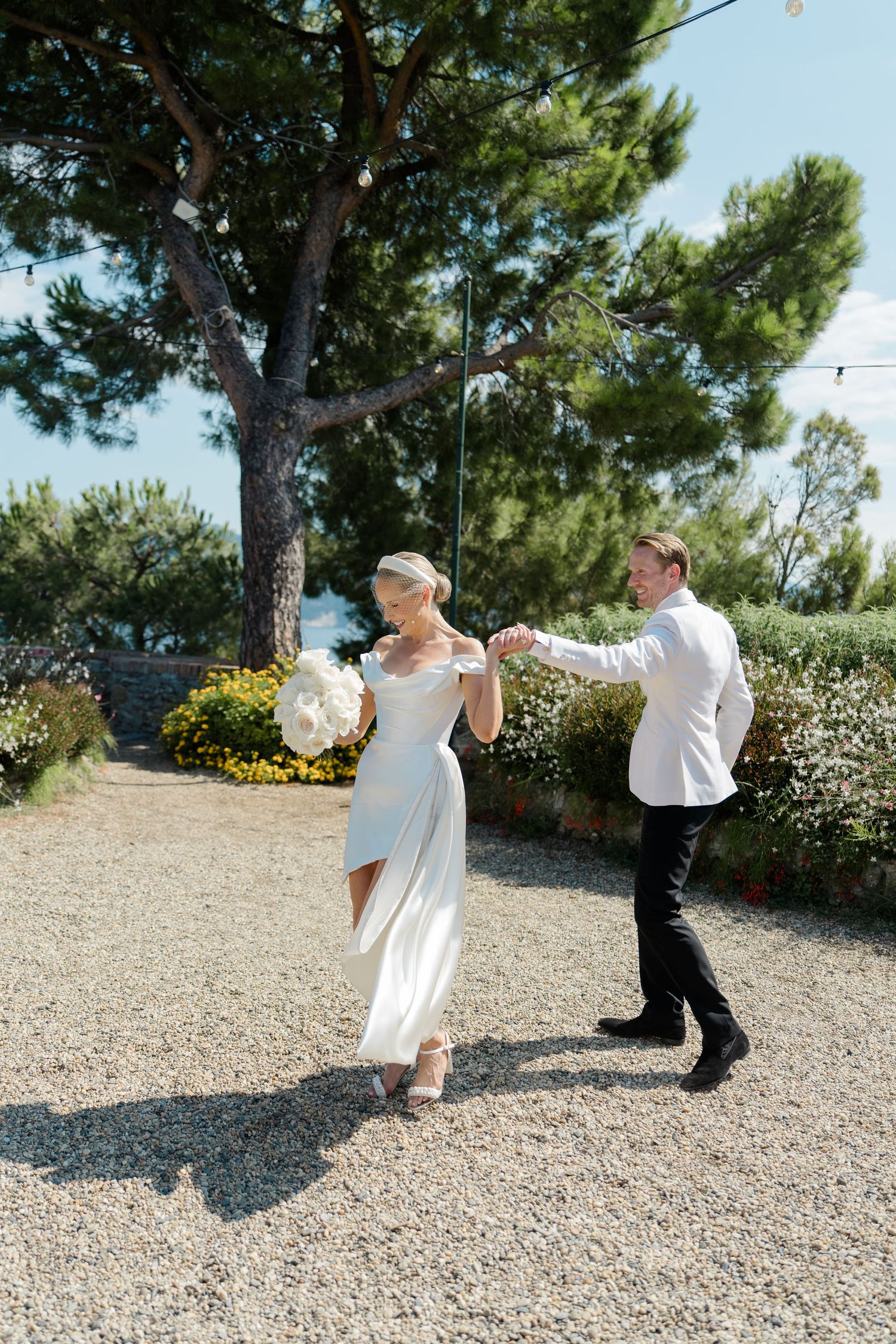 Bride in white gown dances with groom in white jacket on gravel path surrounded by lush garden greenery.
