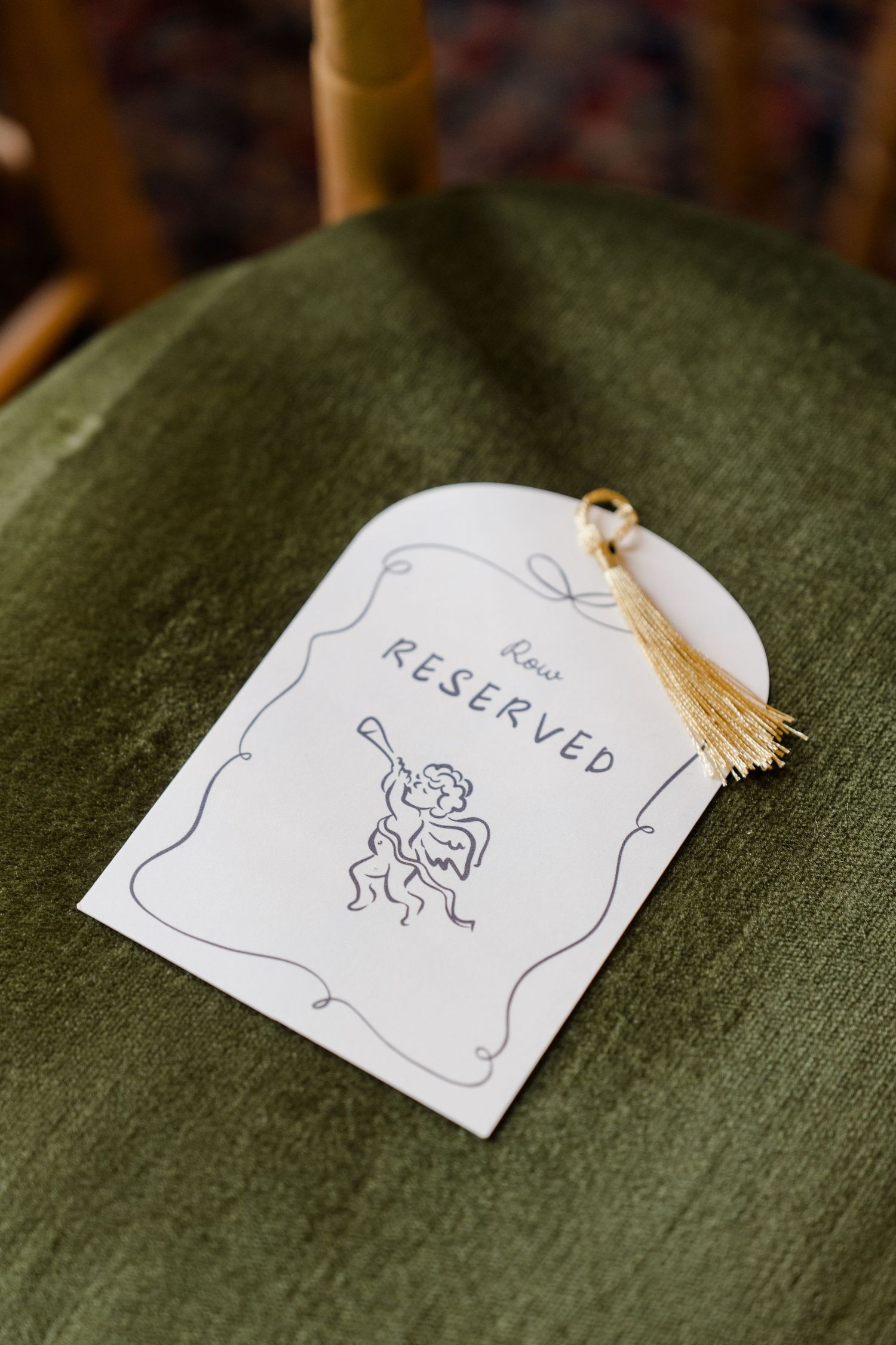 Elegant wedding reserved row card with cherub illustration and gold tassel on green velvet chair.