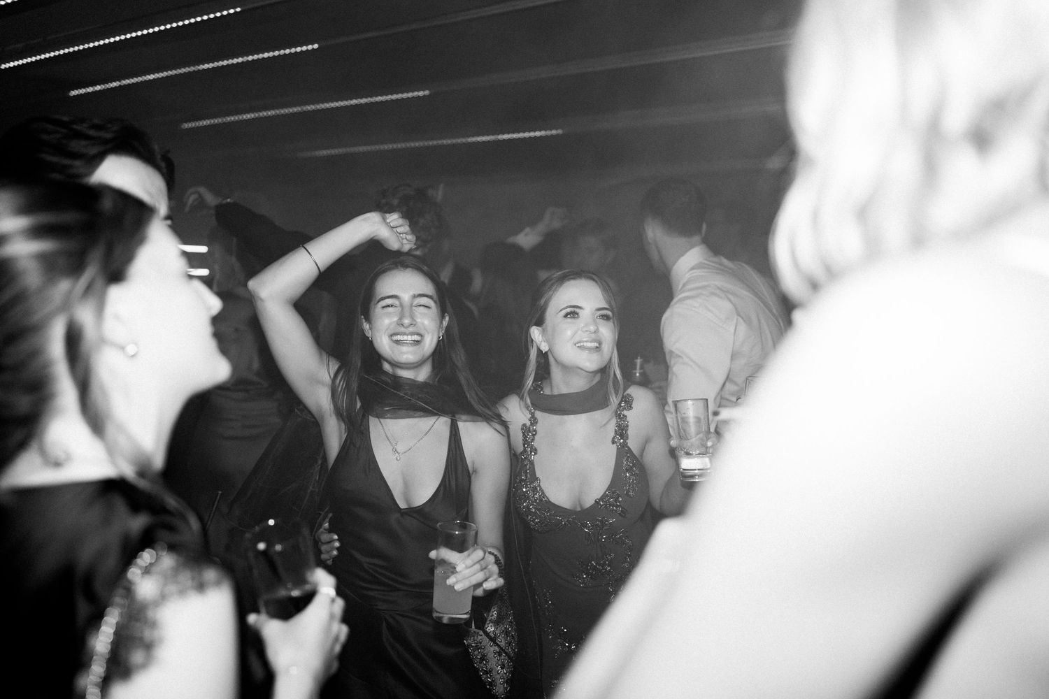 Two women in black dresses dance and smile at a lively wedding reception party.