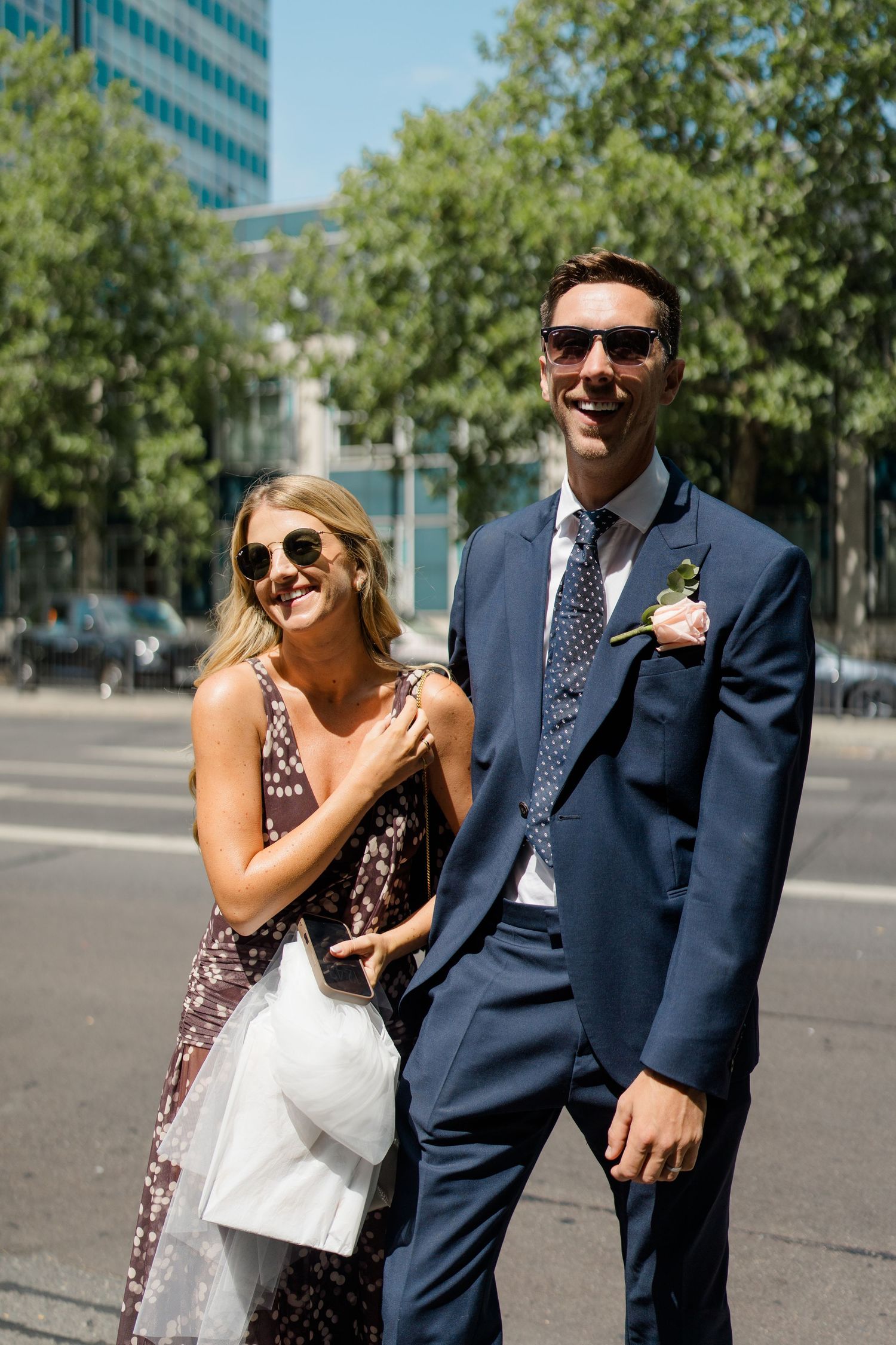 Smiling wedding guests in sunglasses and formal attire pose on sunny city street.