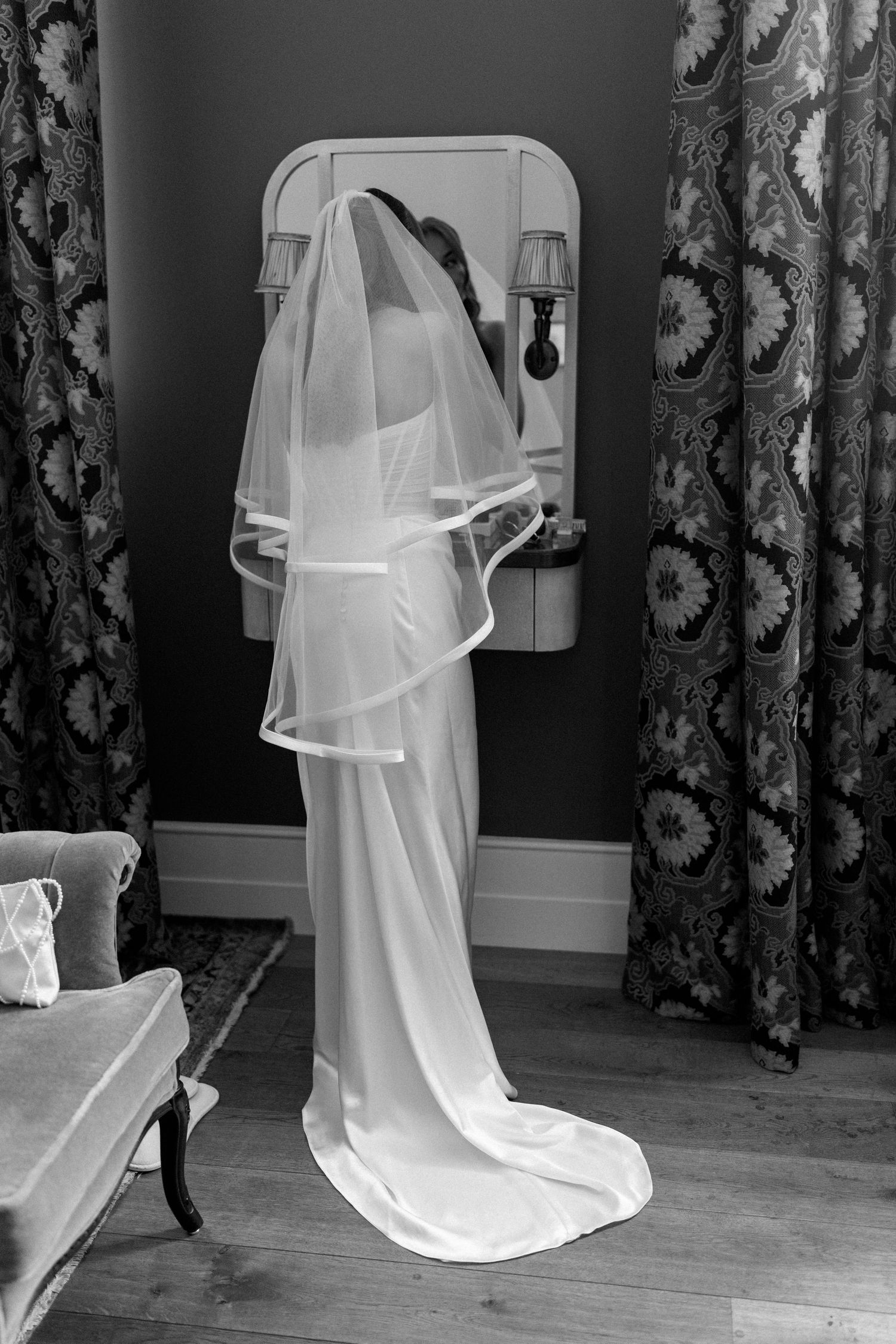 Elegant bride in white wedding dress and veil standing before a mirror in black and white photo.