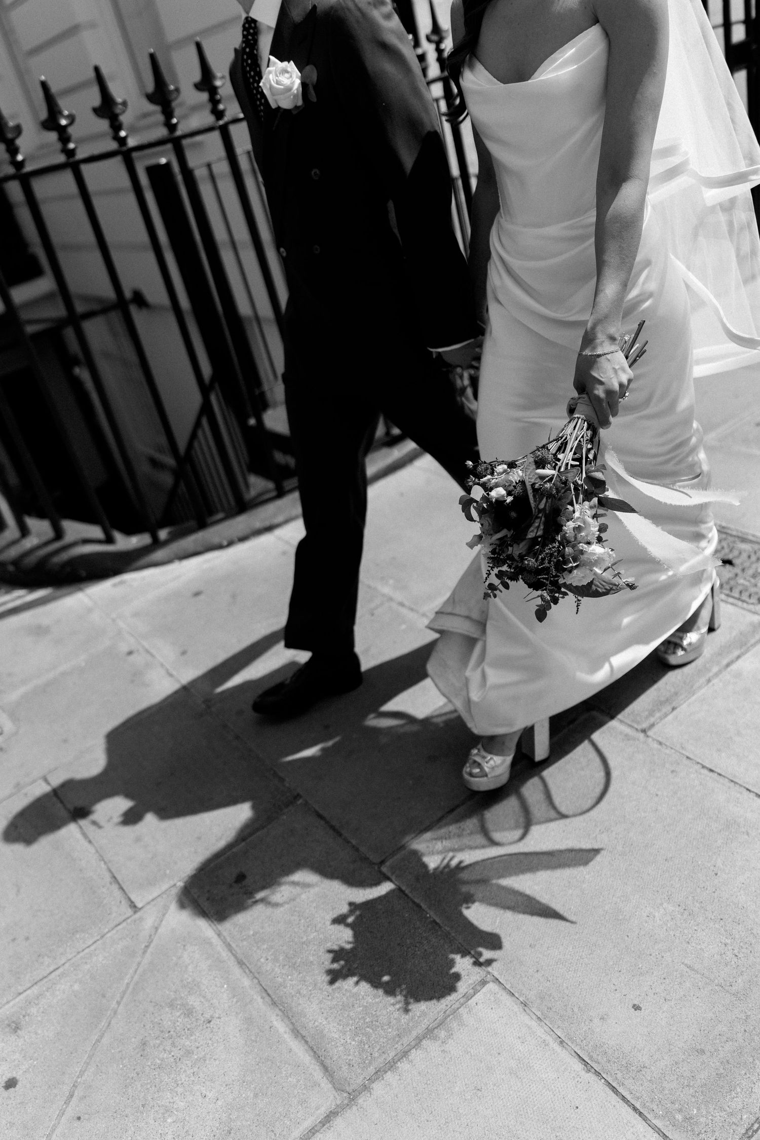 Black and white photo of bride and groom walking on sidewalk, bride holding bouquet with shadows cast on pavement.