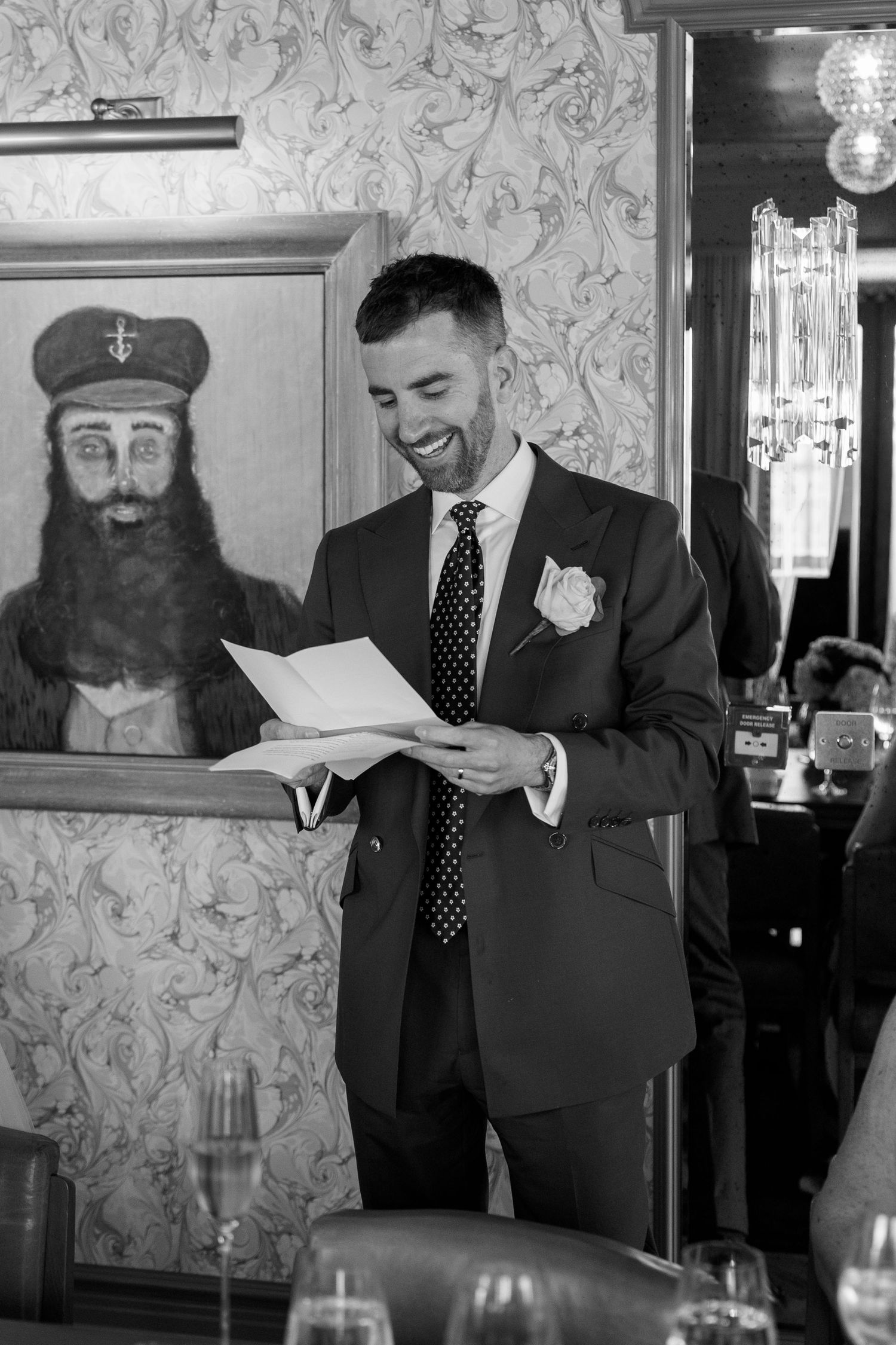 Black and white photo of groom smiling while delivering wedding speech in ornate decorated room.