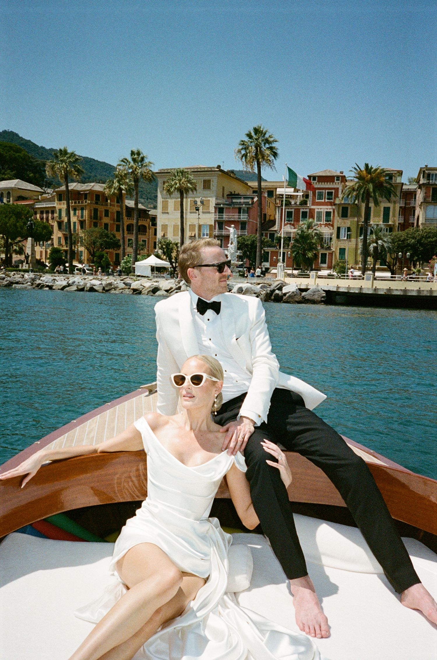 Elegant couple in wedding attire on wooden boat with Italian coastal town in background on sunny day.