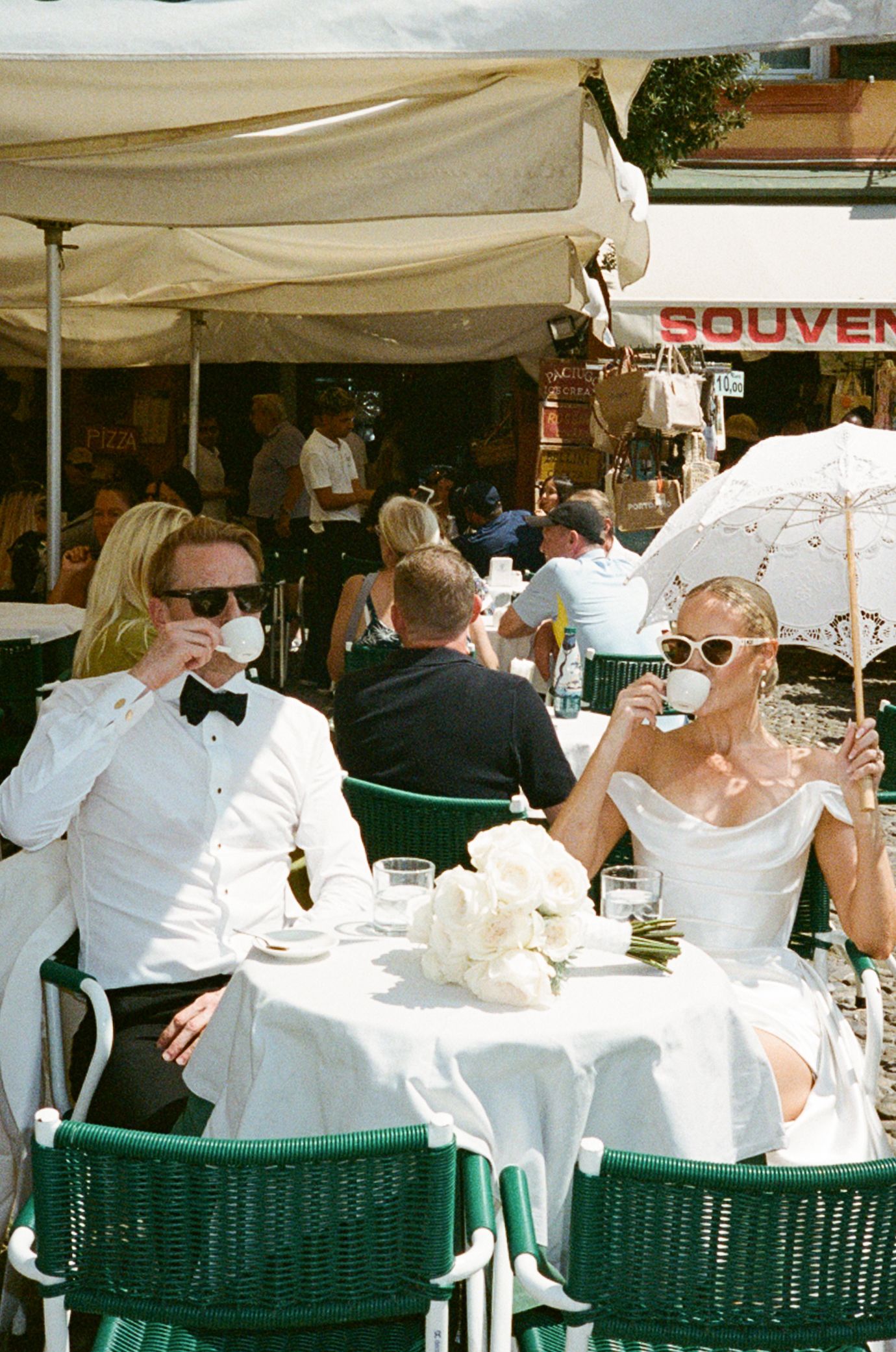 Stylish newlyweds in wedding attire sipping coffee at outdoor Italian cafe terrace on sunny day.