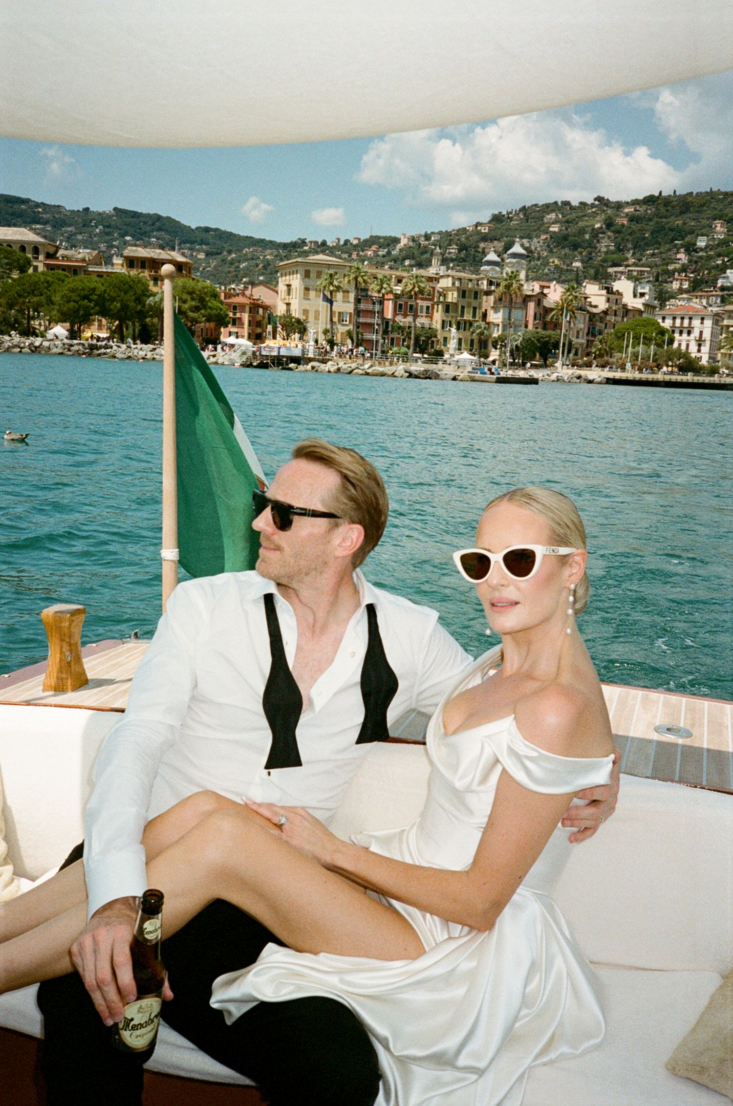 Elegant couple in white outfits and sunglasses relaxing on a luxury boat with Italian coastal town backdrop.