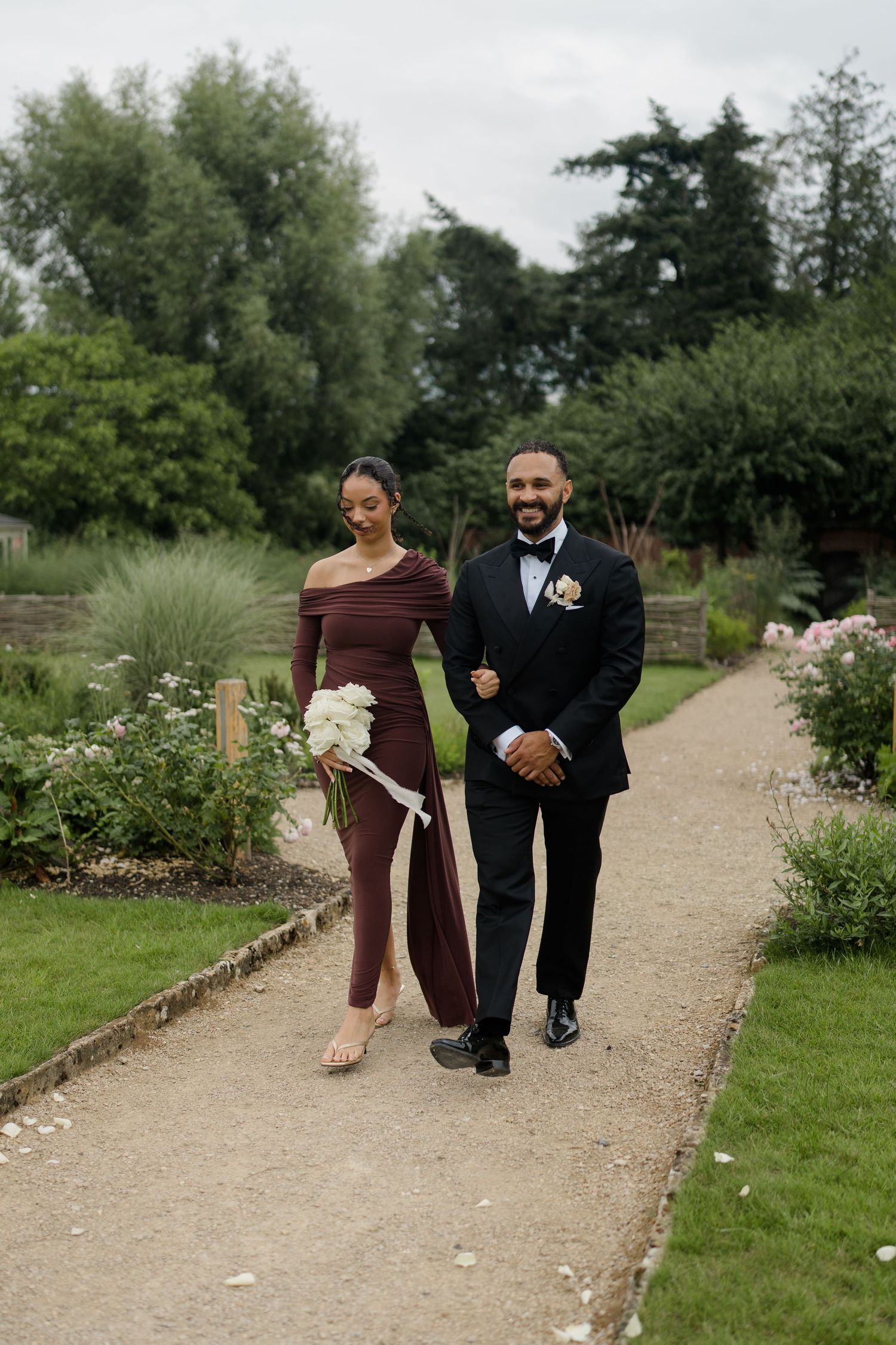 Bridesmaid in burgundy off-shoulder gown and groomsman in black tuxedo walk together along garden wedding pathway.