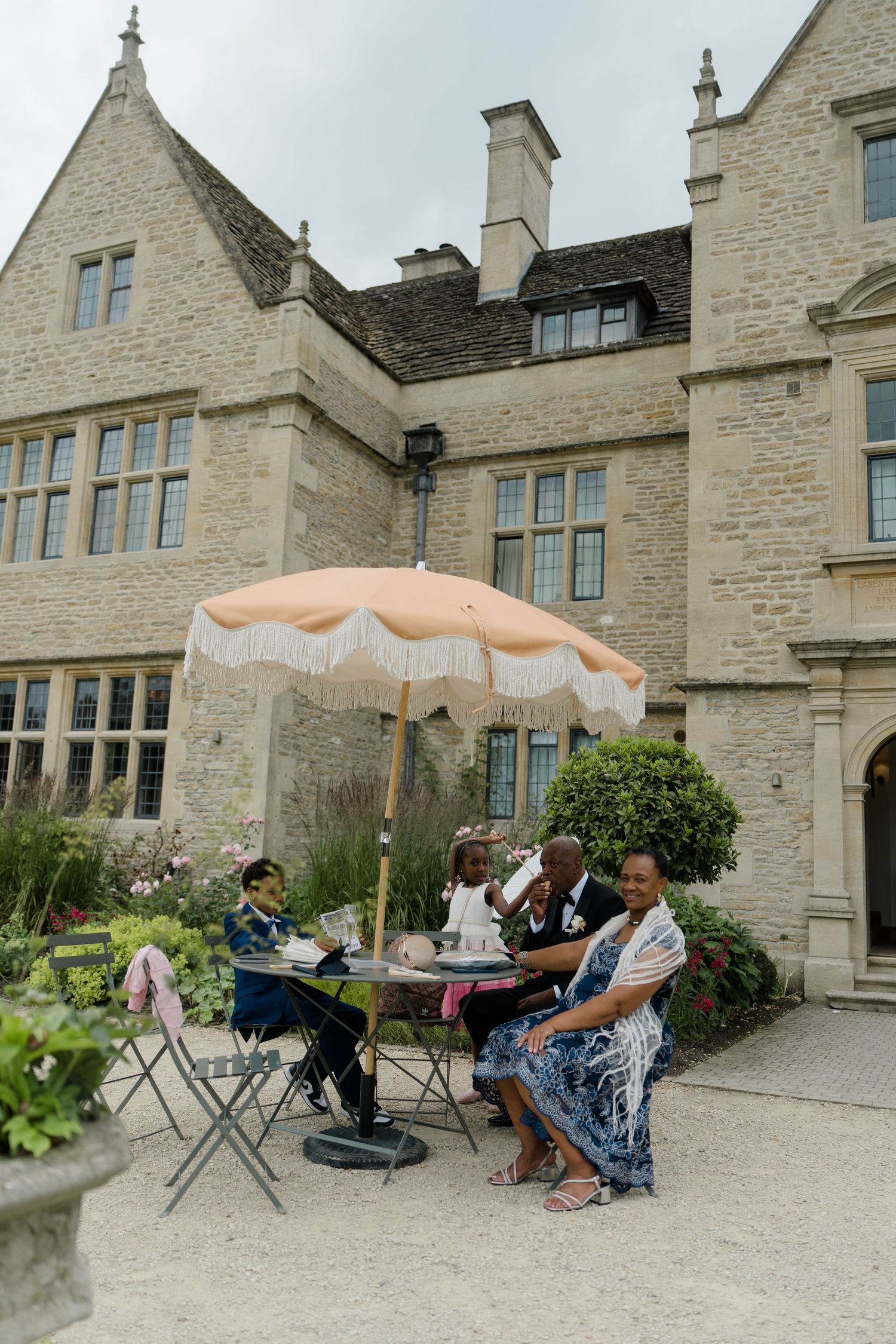 Wedding guests relaxing under fringed parasol at outdoor table beside historic stone manor.