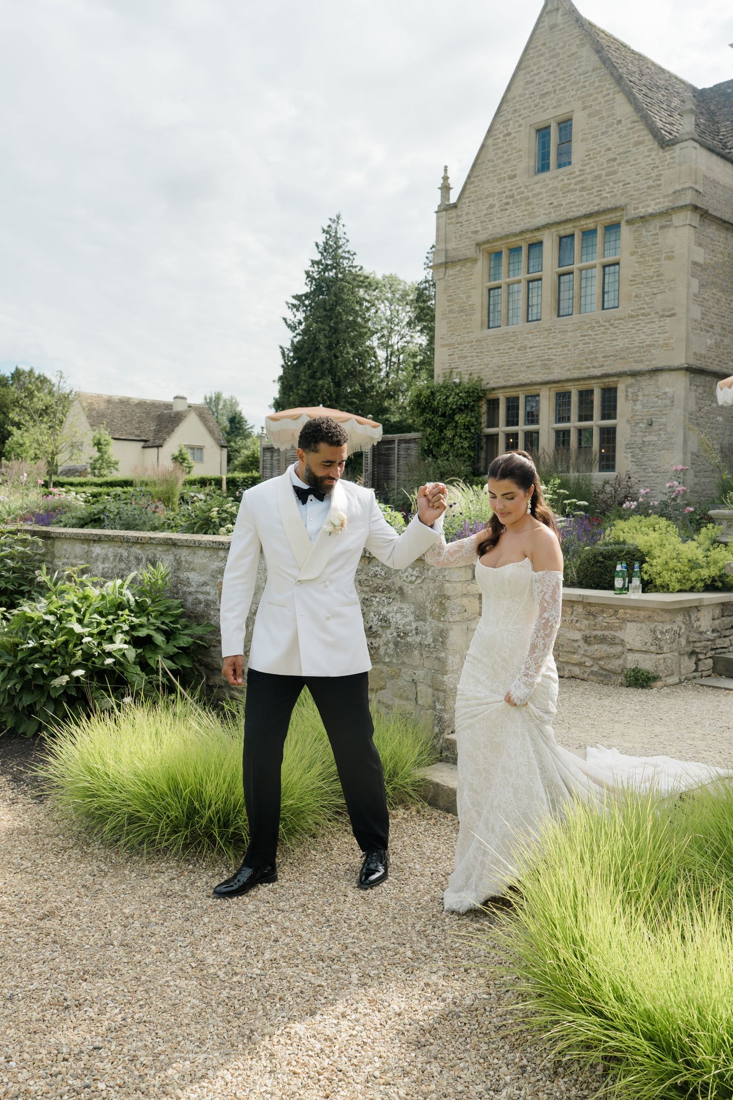 Groom in white tuxedo jacket holds brides hand in lace gown at elegant English countryside wedding.