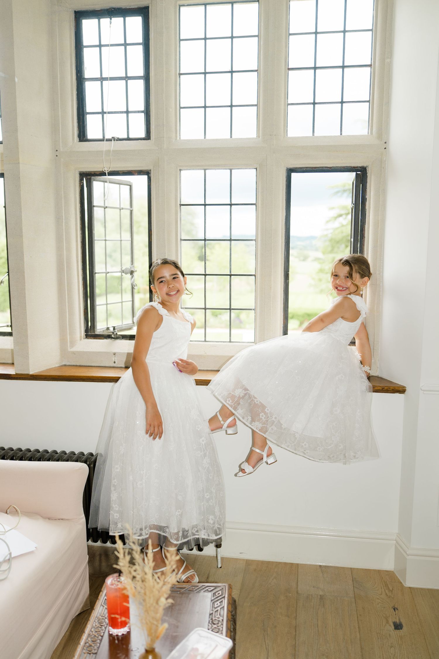 Two flower girls in white lace dresses playing near large windows in a bright elegant room.