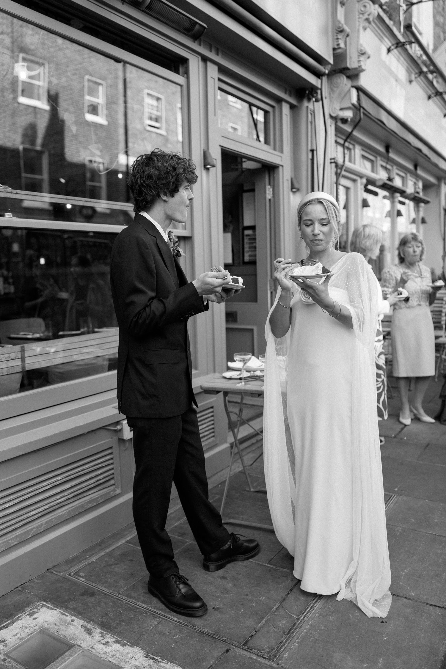 A couple in wedding attire shares cake outside their reception venue in candid shots.