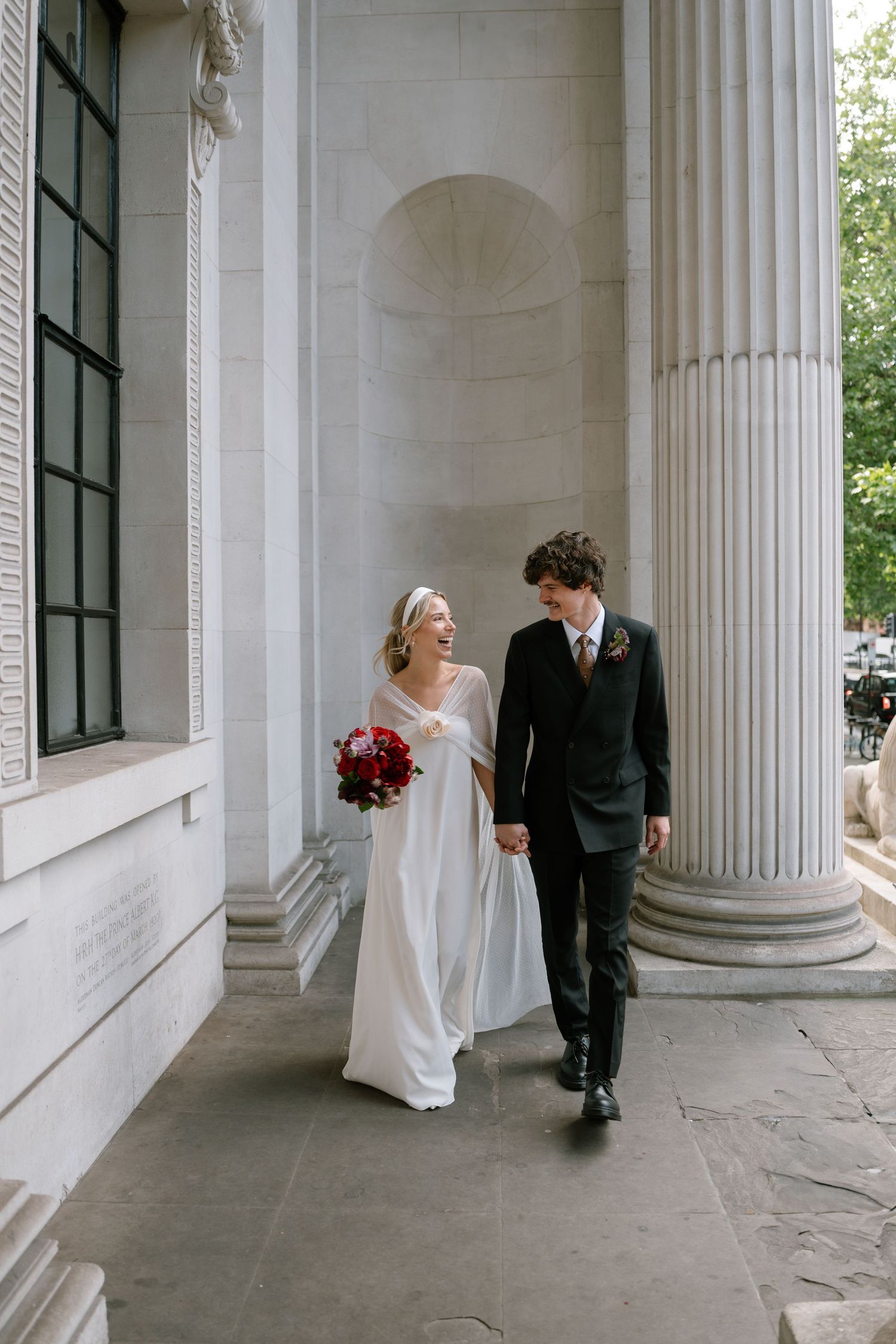 Newlyweds walking hand in hand along columned corridor of historic building.