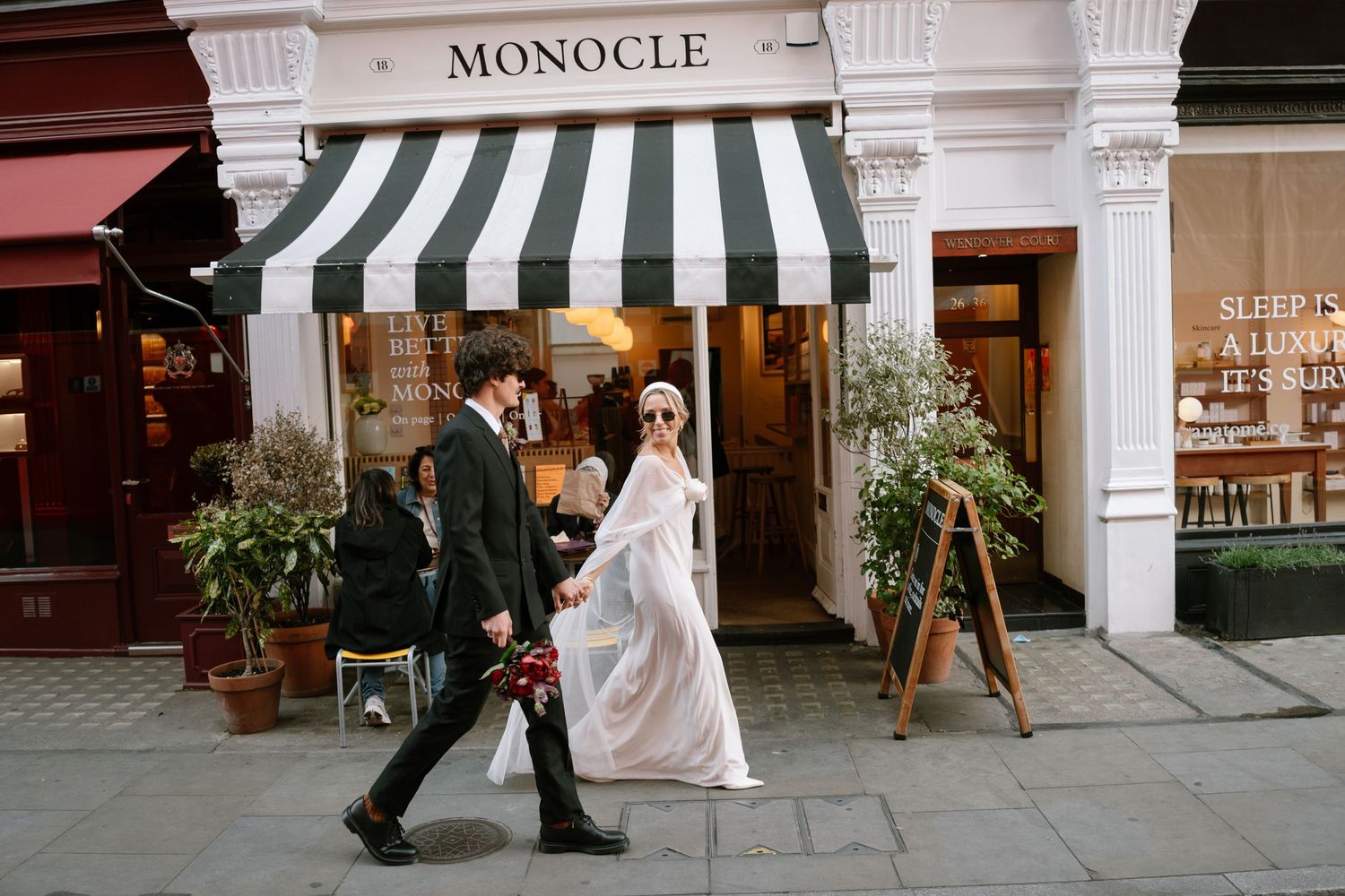Wedding couple strolling past elegant storefronts with white columns and striped awnings.