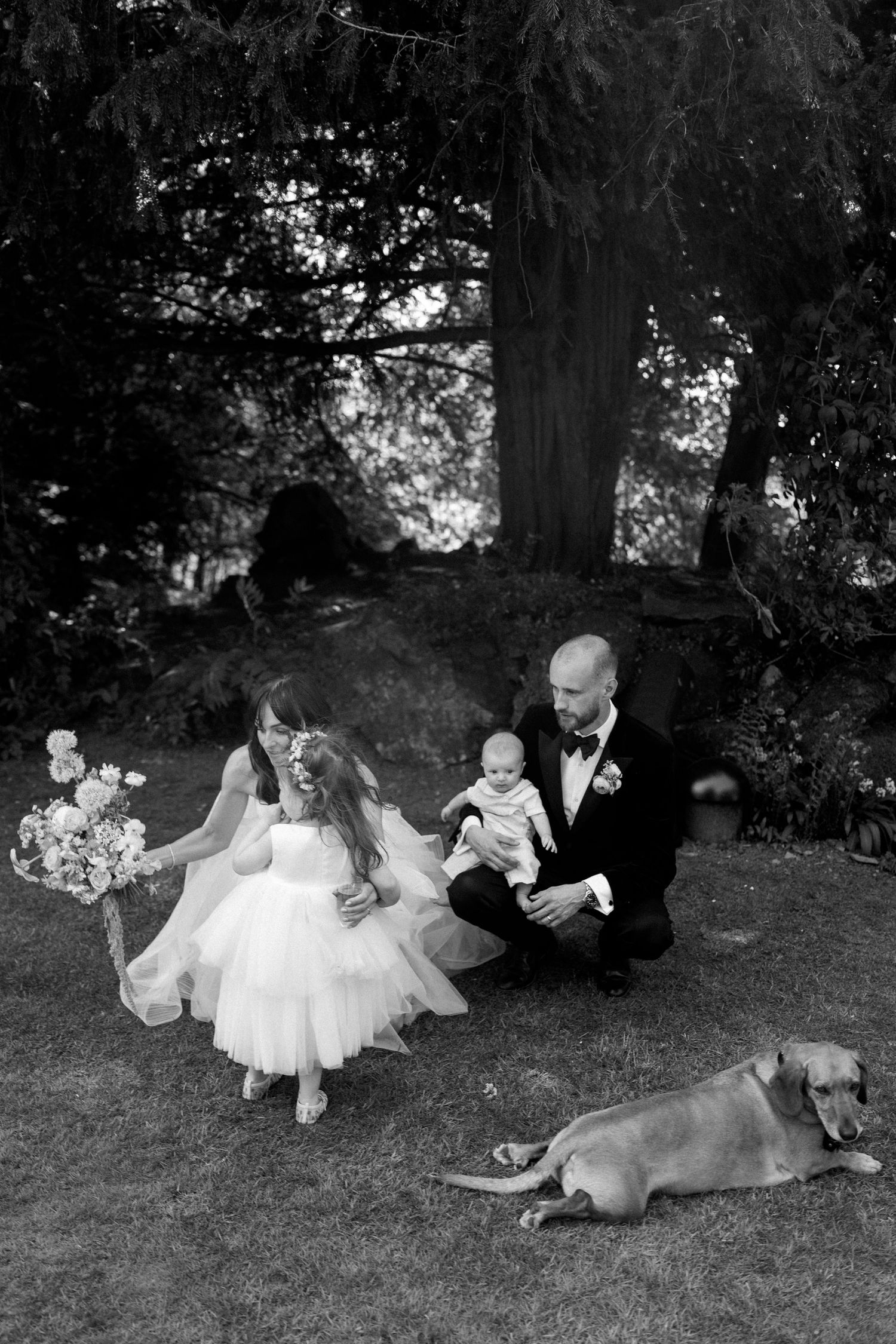 A family sits on grass with their dog during a wedding celebration in a garden setting.