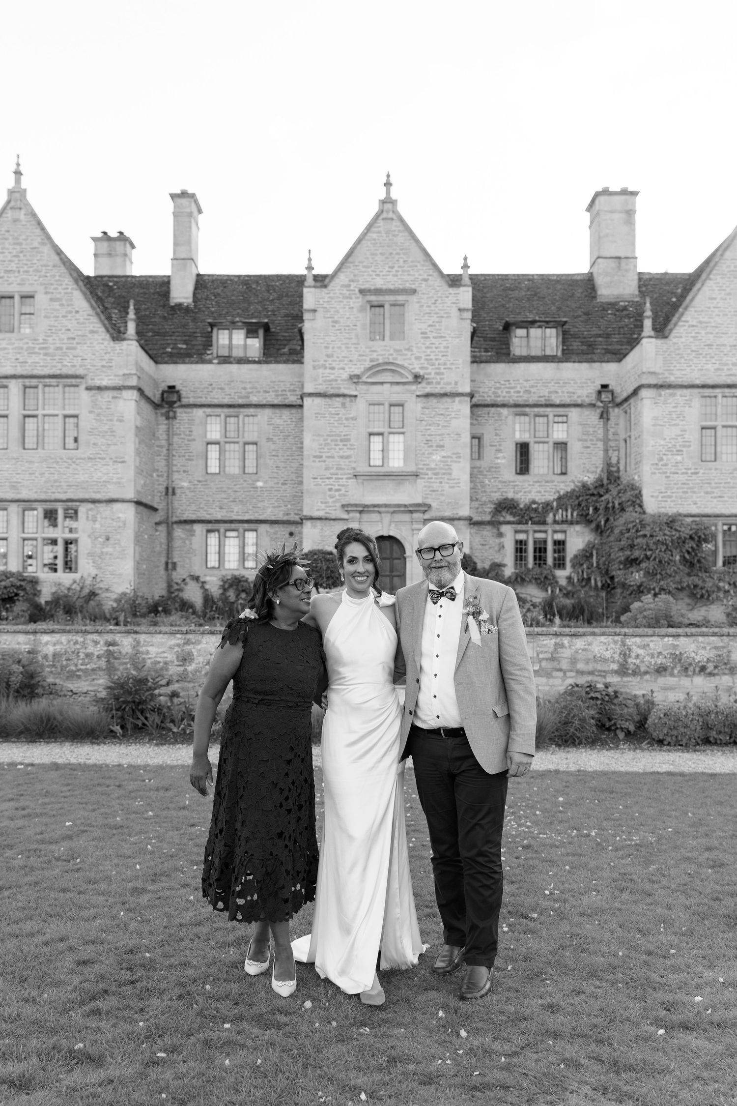 Black and white photo of bride in halter gown posing with two guests in front of grand stone manor house.