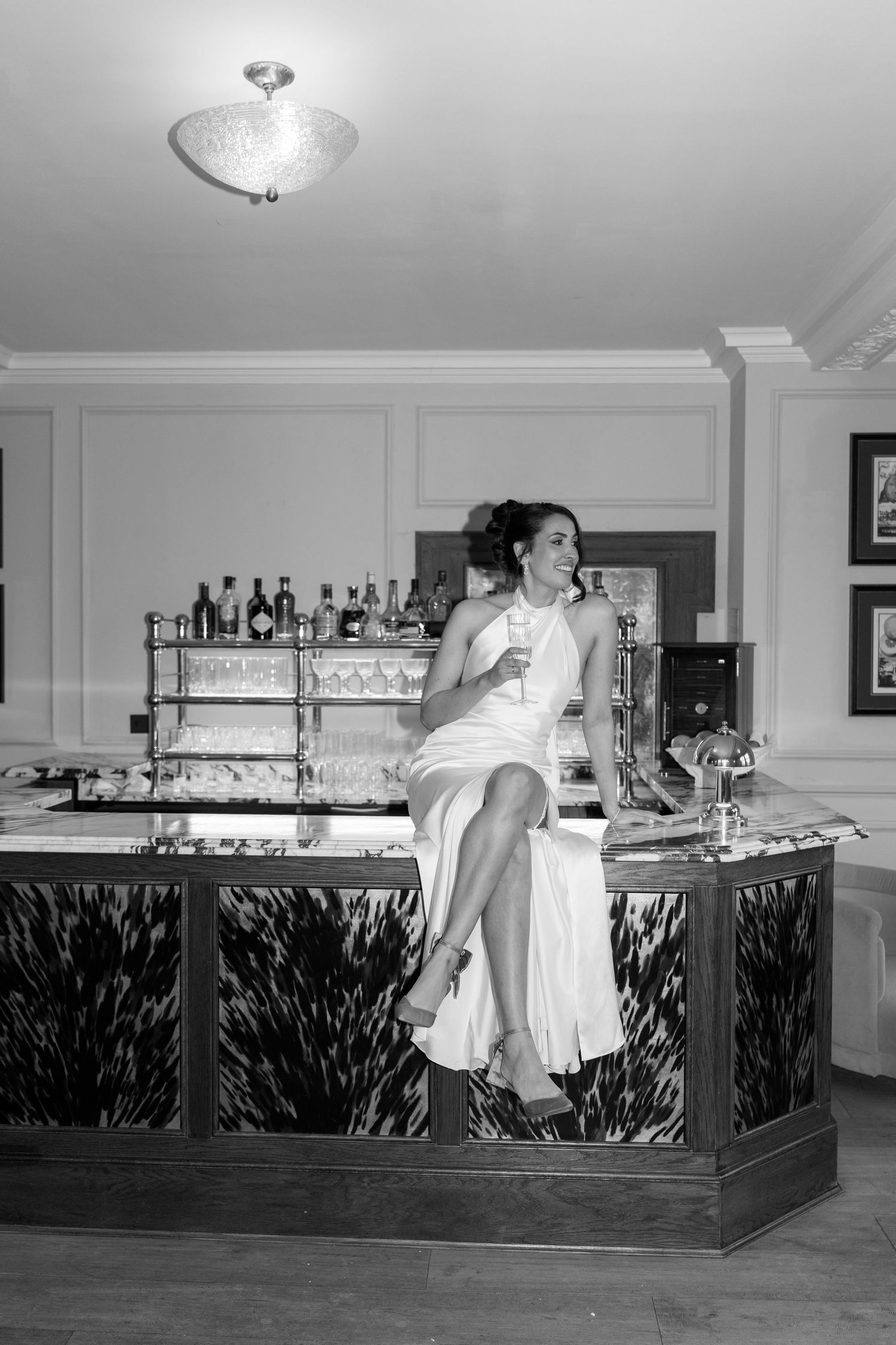 Black and white photo of bride sitting on bar counter holding champagne glass in elegant venue.