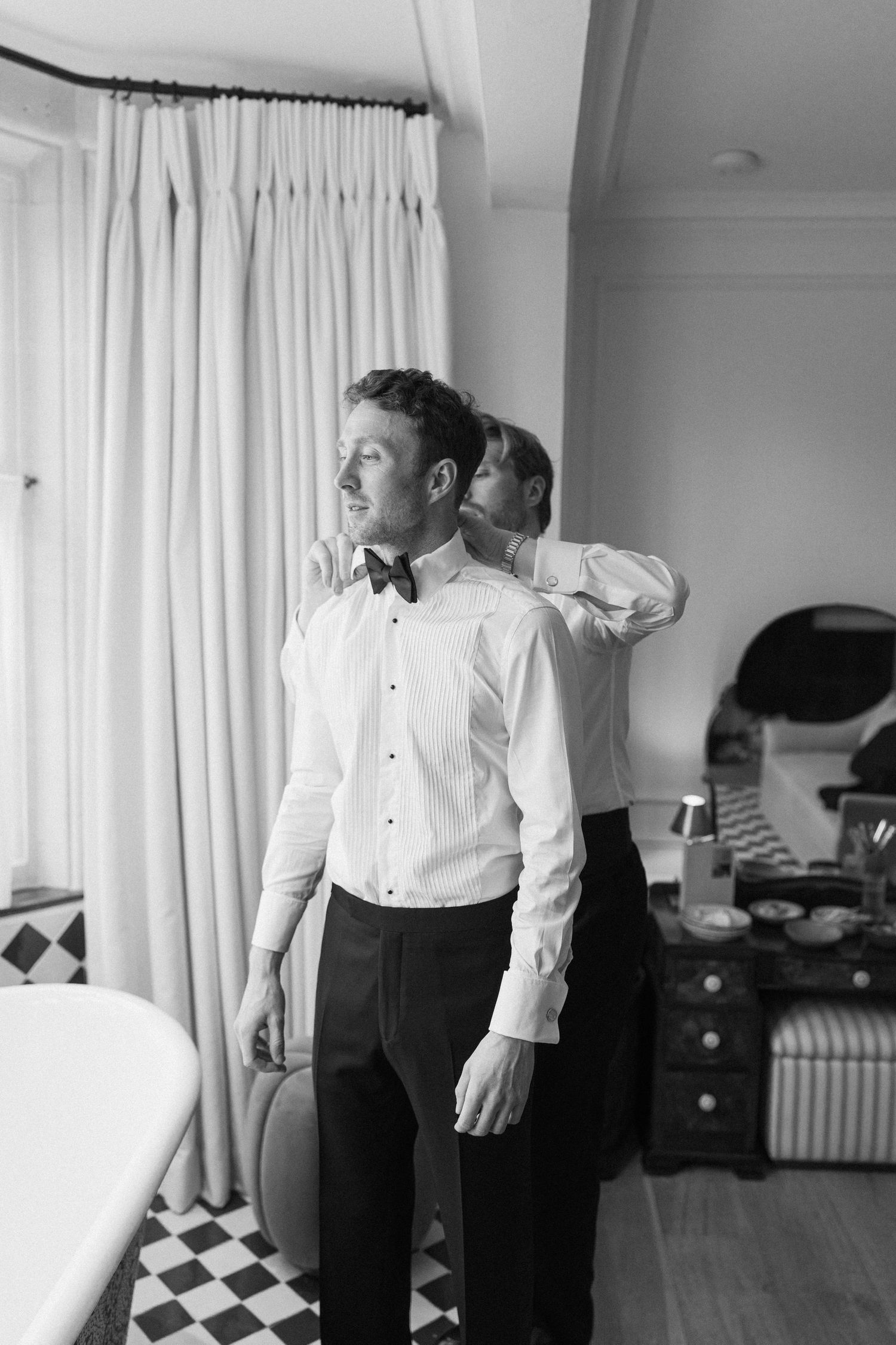 Black and white photo of groom in white shirt and bow tie being helped to dress by groomsman.