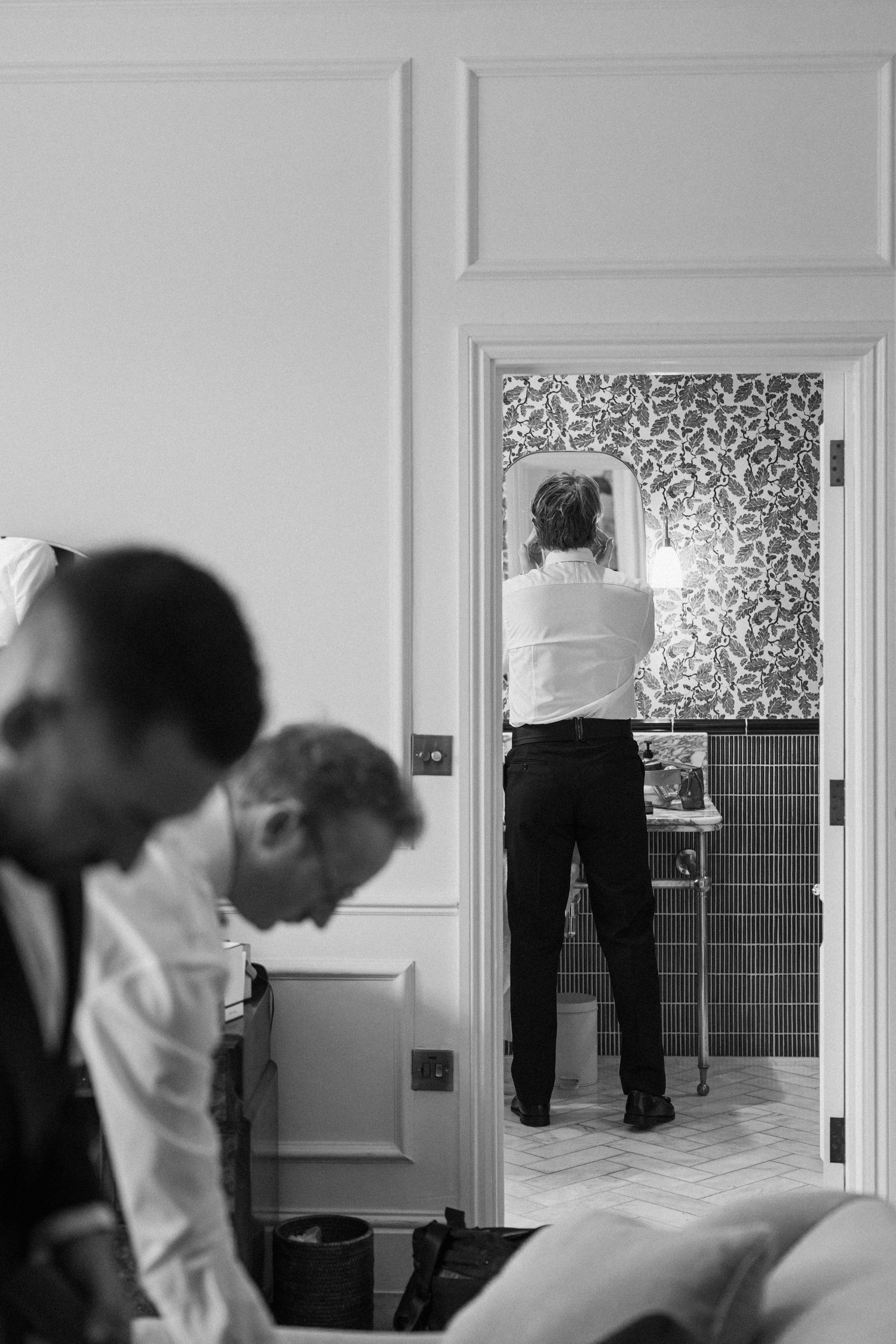 Groom preparing in bathroom mirror while groomsmen get ready in black and white wedding photo.
