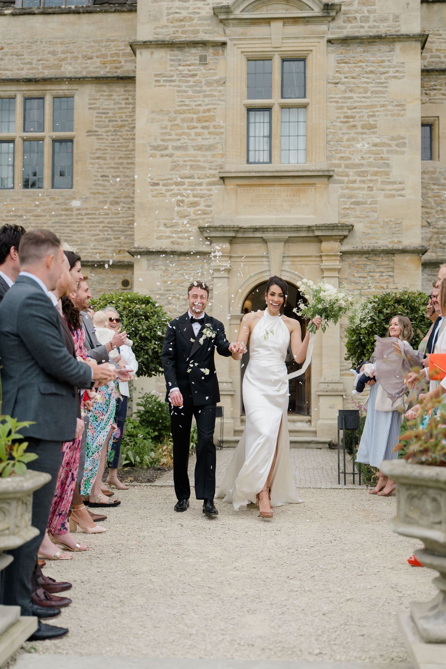 Bride and groom walk through confetti shower outside stone manor wedding venue with guests lining the path.