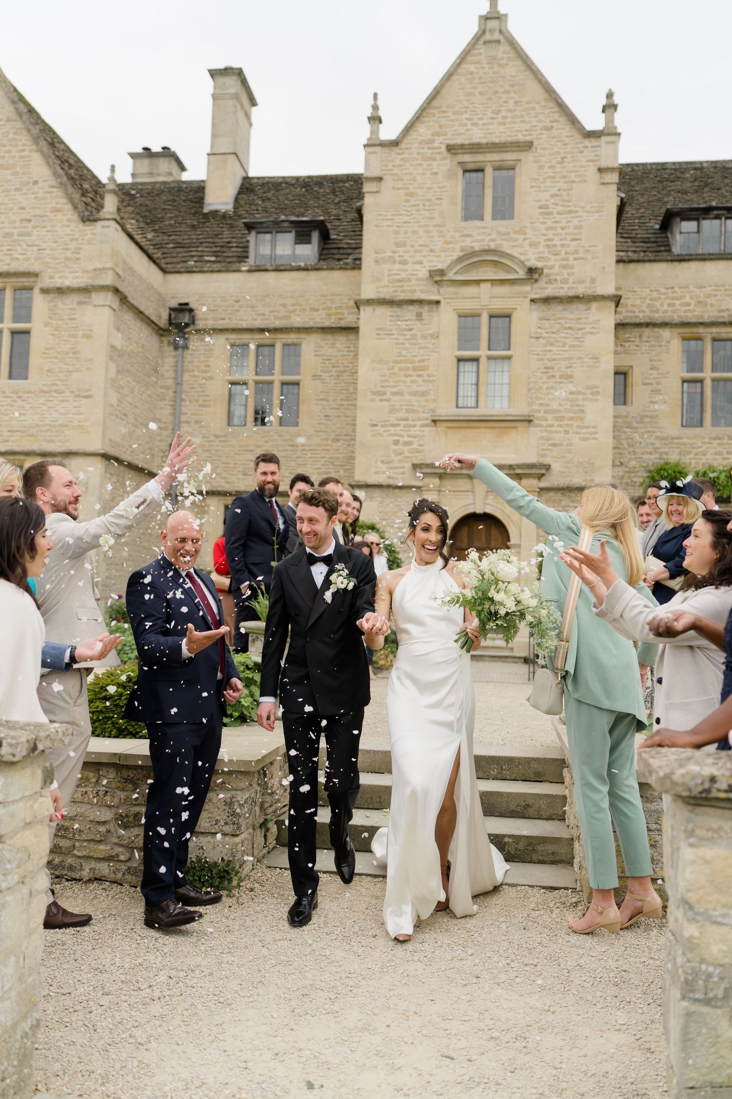 Bride and groom walk through confetti shower outside a stunning Cotswolds stone manor house surrounded by cheering wedding guests.