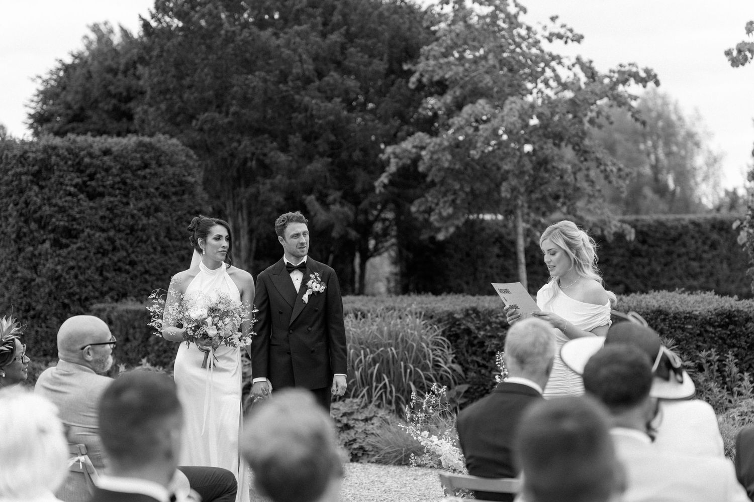 A couple stands at an outdoor wedding ceremony while an officiant reads from a paper.