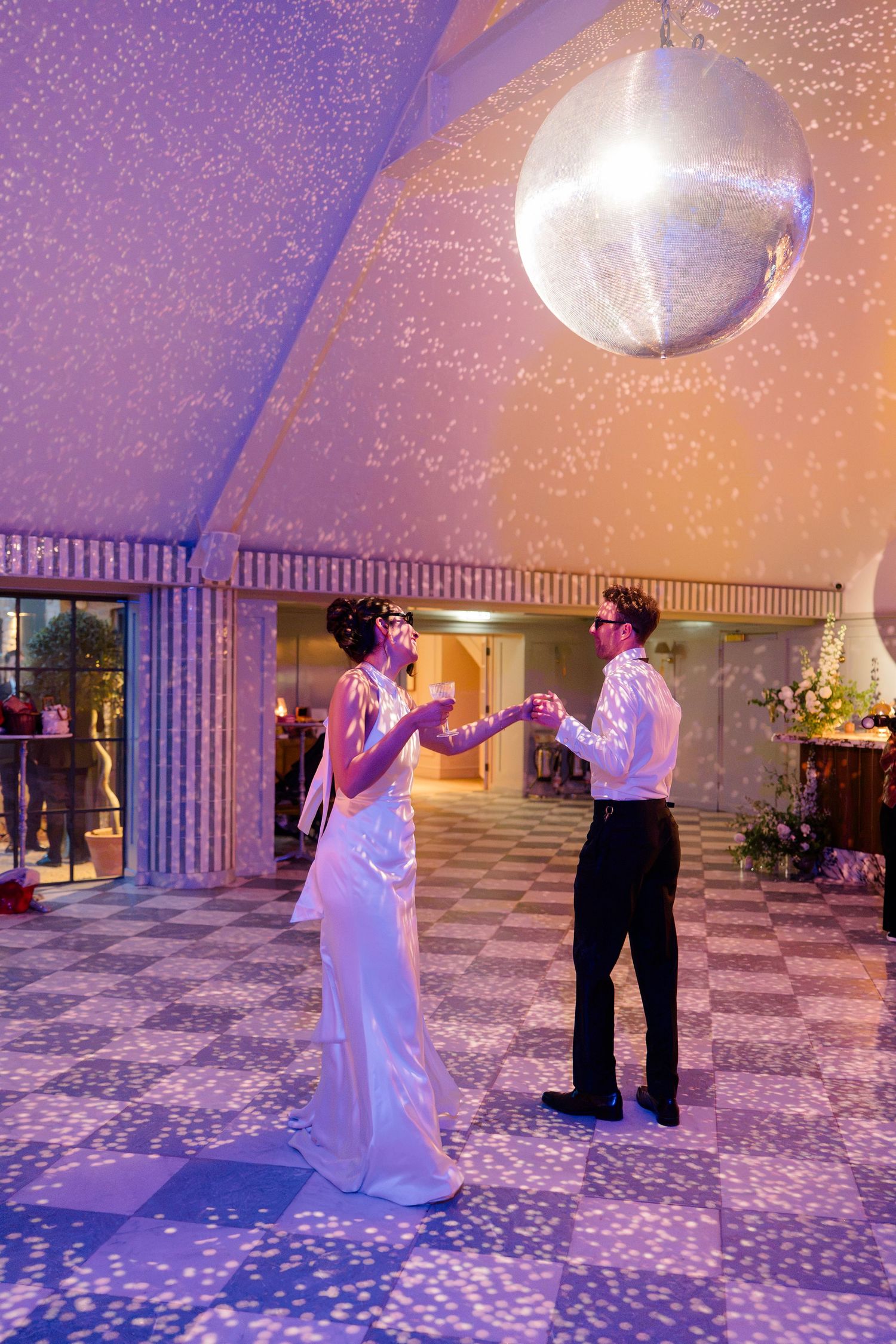 Bride and groom share first dance under a glittering disco ball in a purple-lit venue.