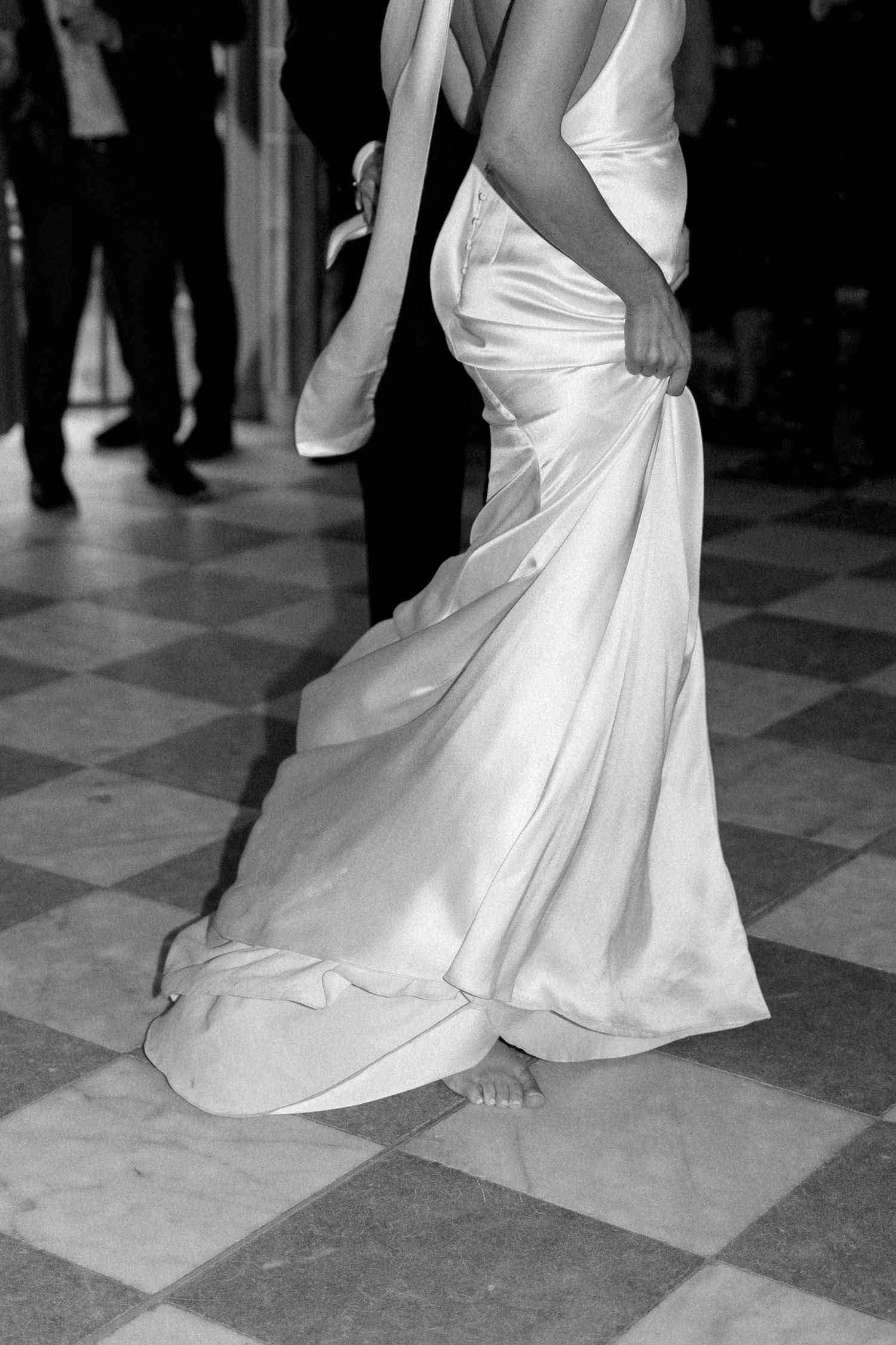 Black and white close-up of bride dancing barefoot in flowing satin wedding gown on checkered floor.