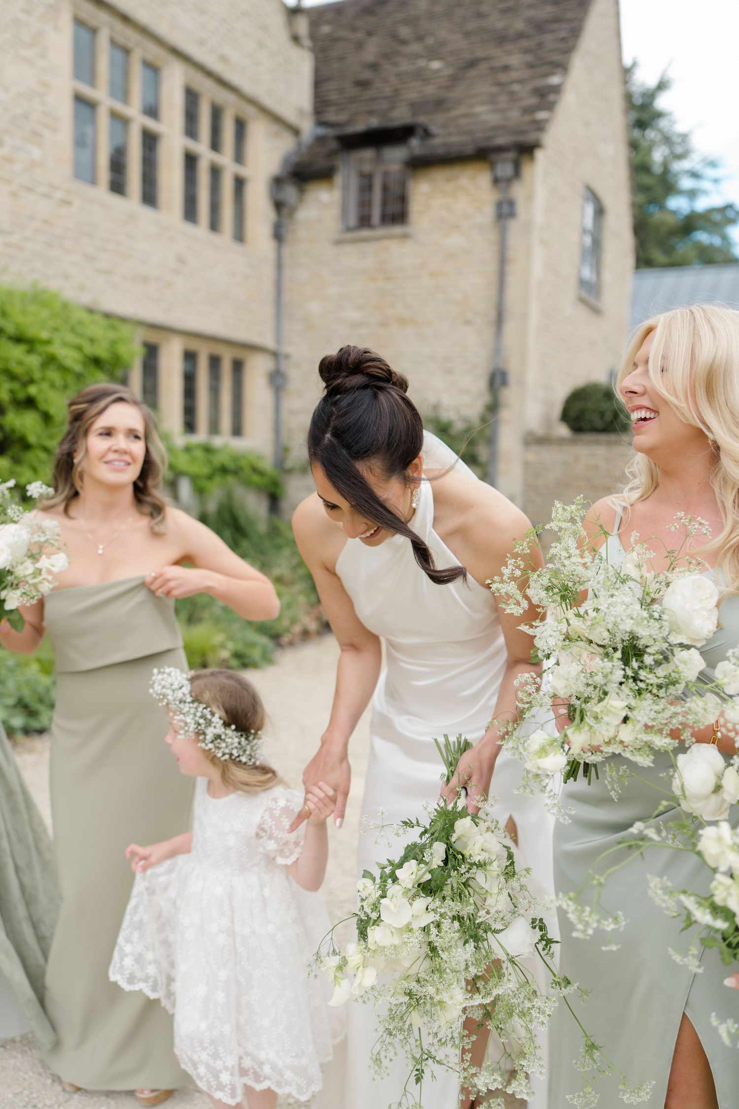 Bride in white halter gown walks with bridesmaids in sage dresses holding white floral bouquets.