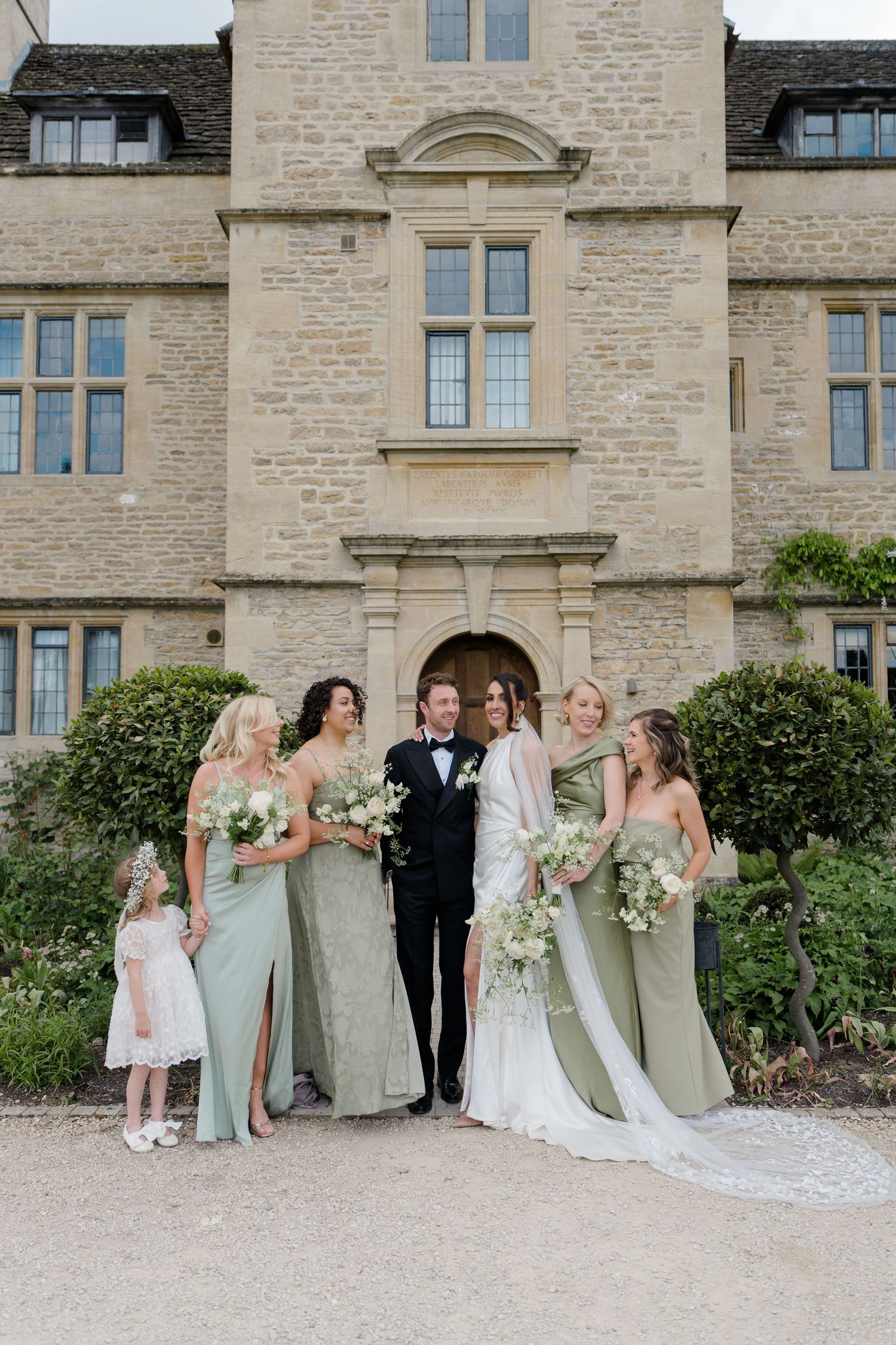 Wedding party with bride, groom, bridesmaids in sage green gowns and flower girl posing before historic stone manor.