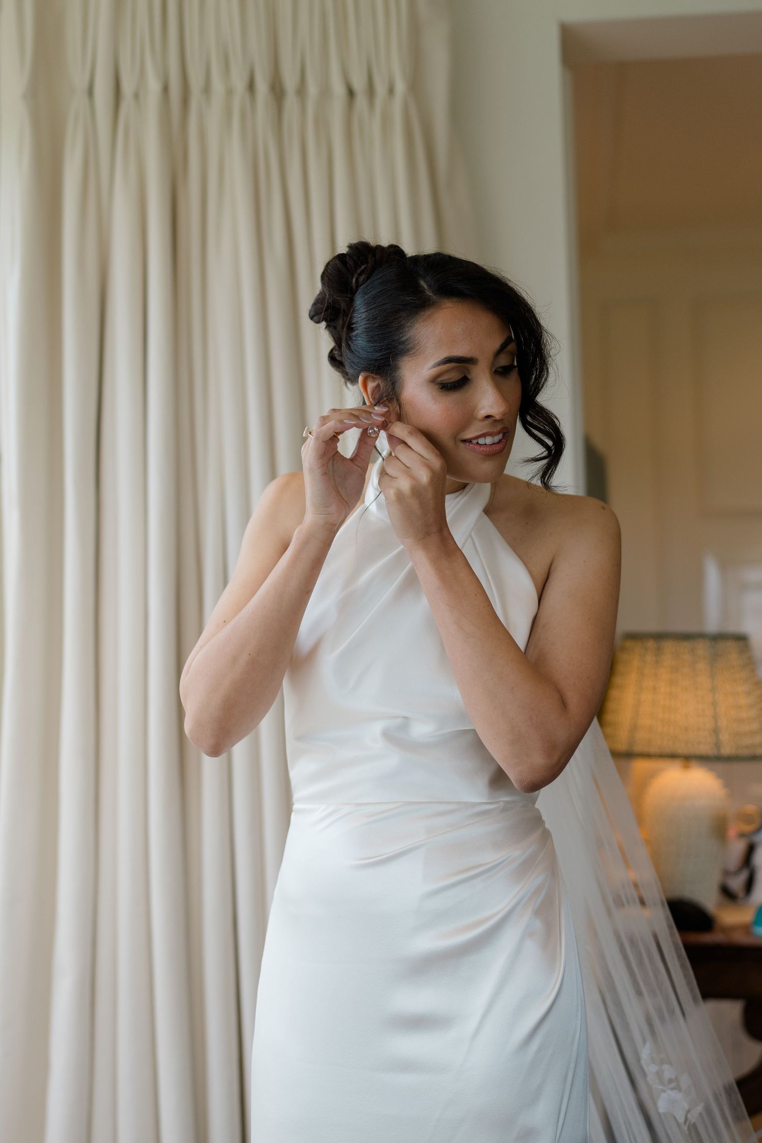 Elegant bride in white halter gown putting on earrings while smiling in softly lit room.