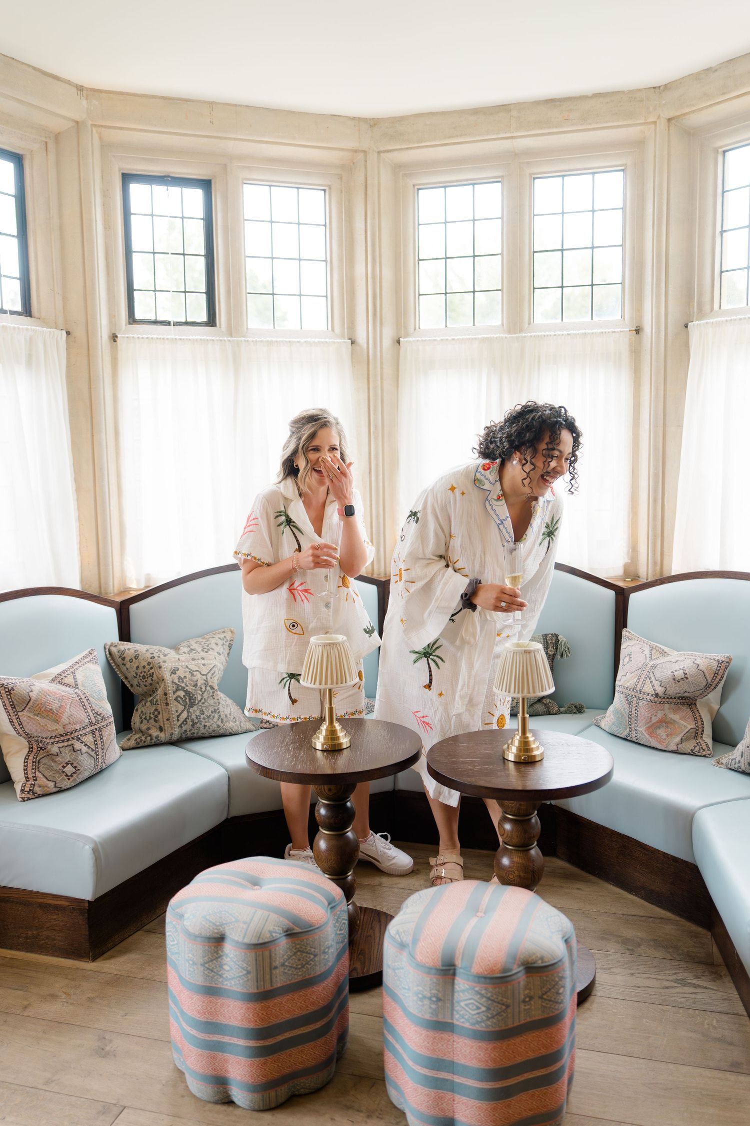 Two women in matching robes enjoying drinks in a bright bay window sitting room with blue upholstered curved seating.