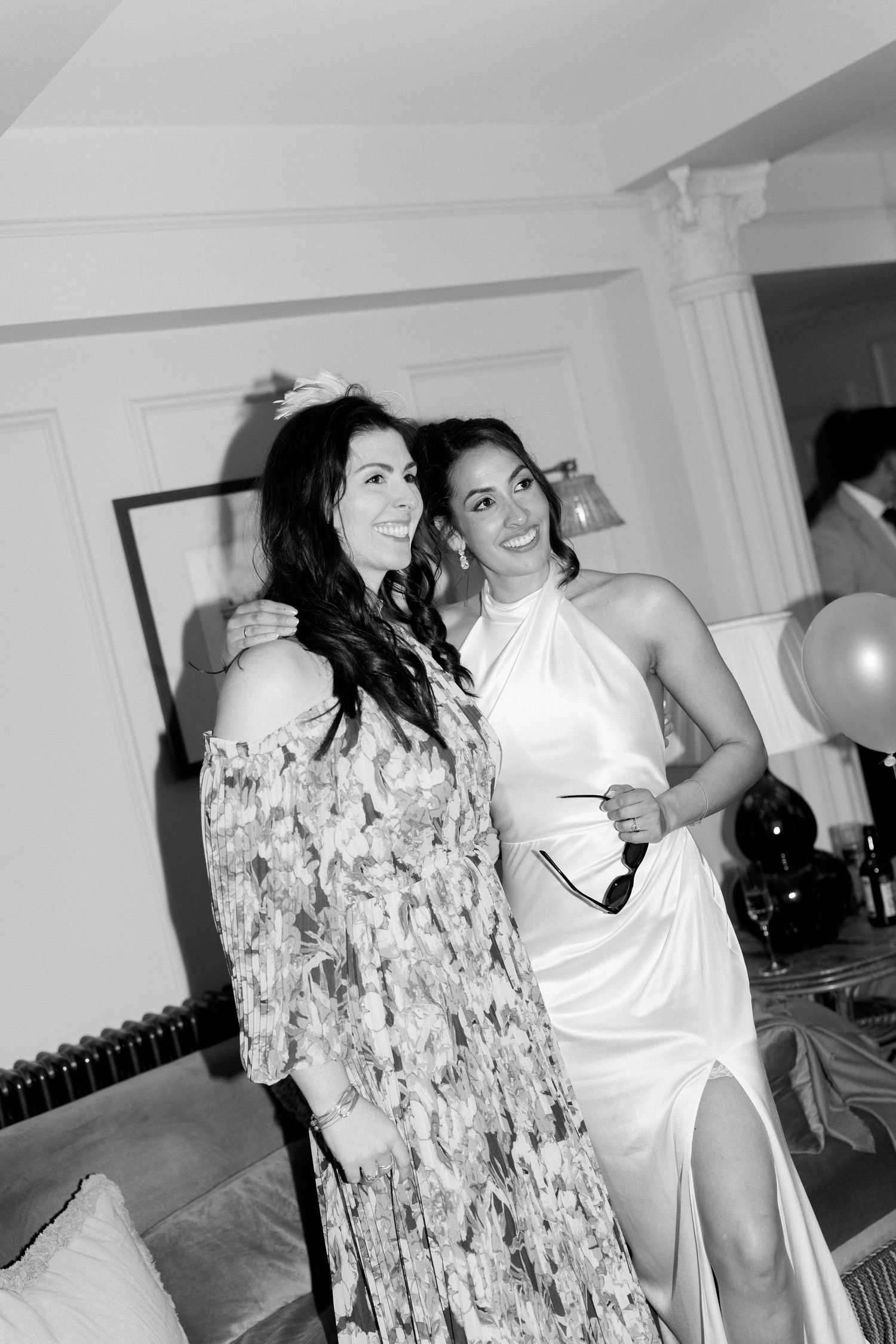 Bride in white halter gown smiles with a friend in floral dress at a wedding celebration.