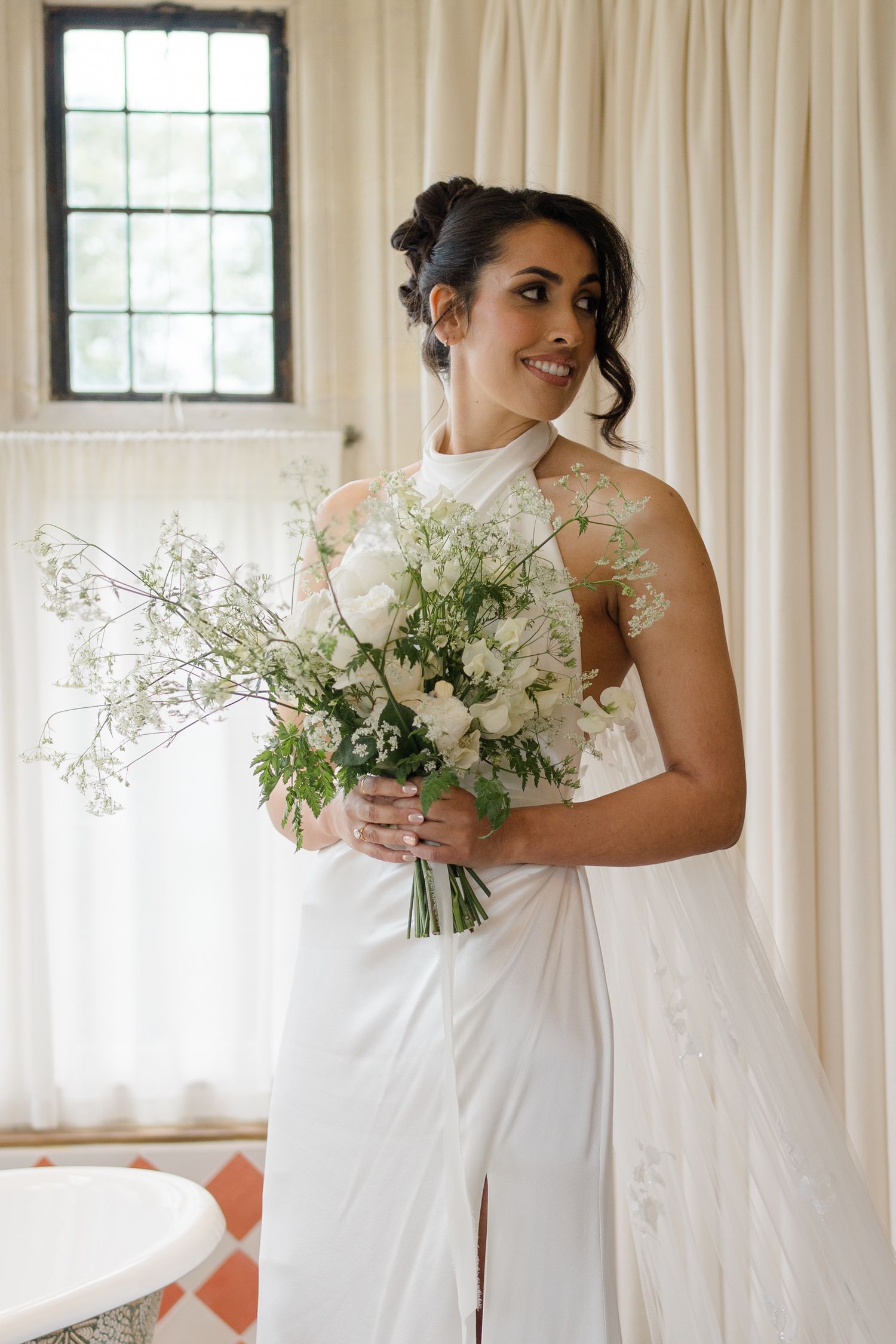 Radiant bride in white halter gown holding a lush white and green bouquet in a bright, airy room.