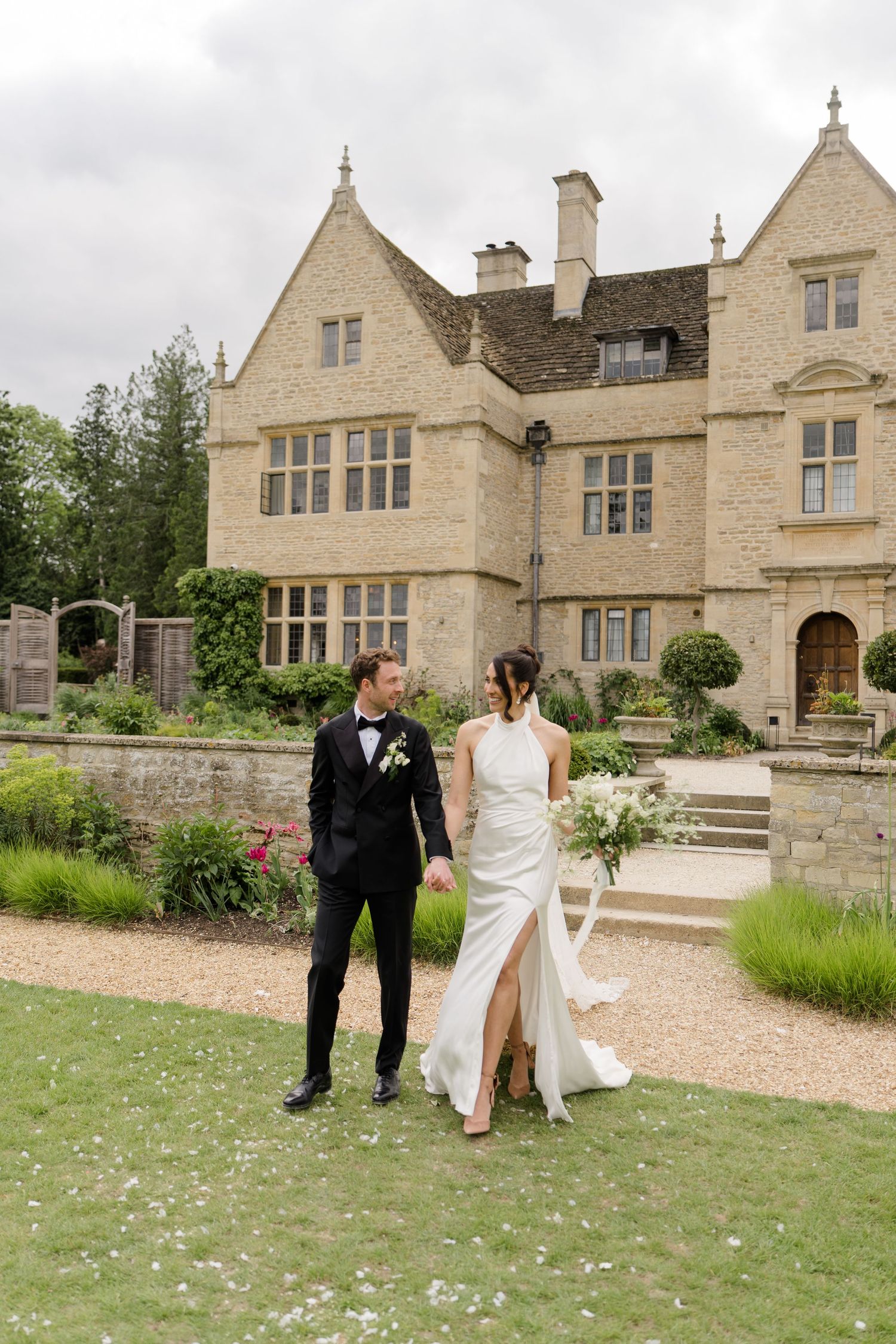 Bride and groom hold hands walking in garden of elegant stone English country house venue.