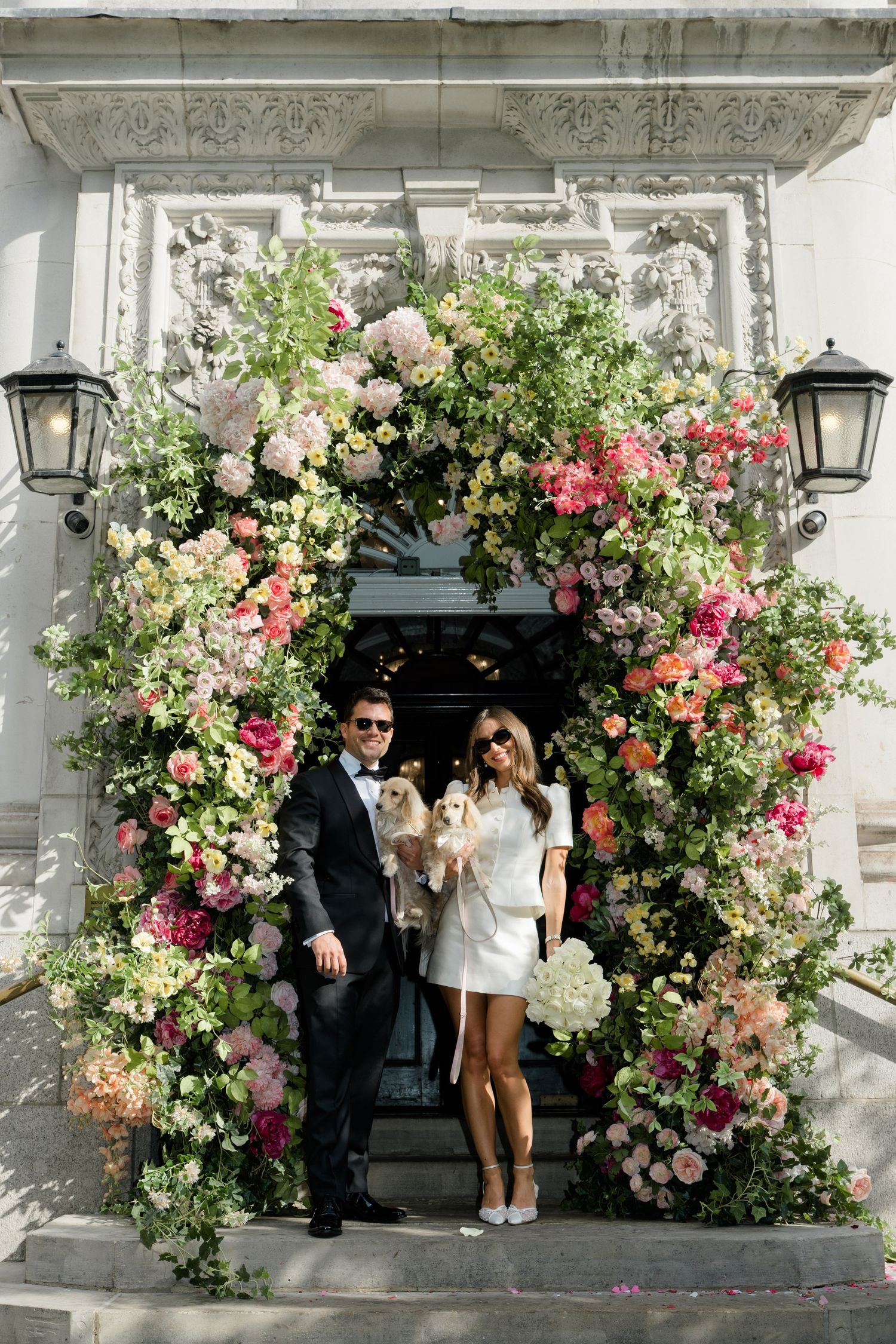 Couple in wedding attire hold dogs under stunning floral arch with colorful blooms at venue.