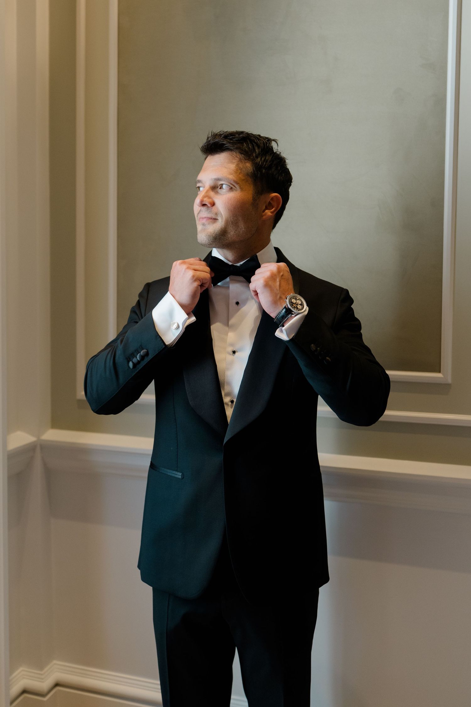 Dapper groom in black tuxedo adjusting bow tie while smiling in an elegant hotel room.