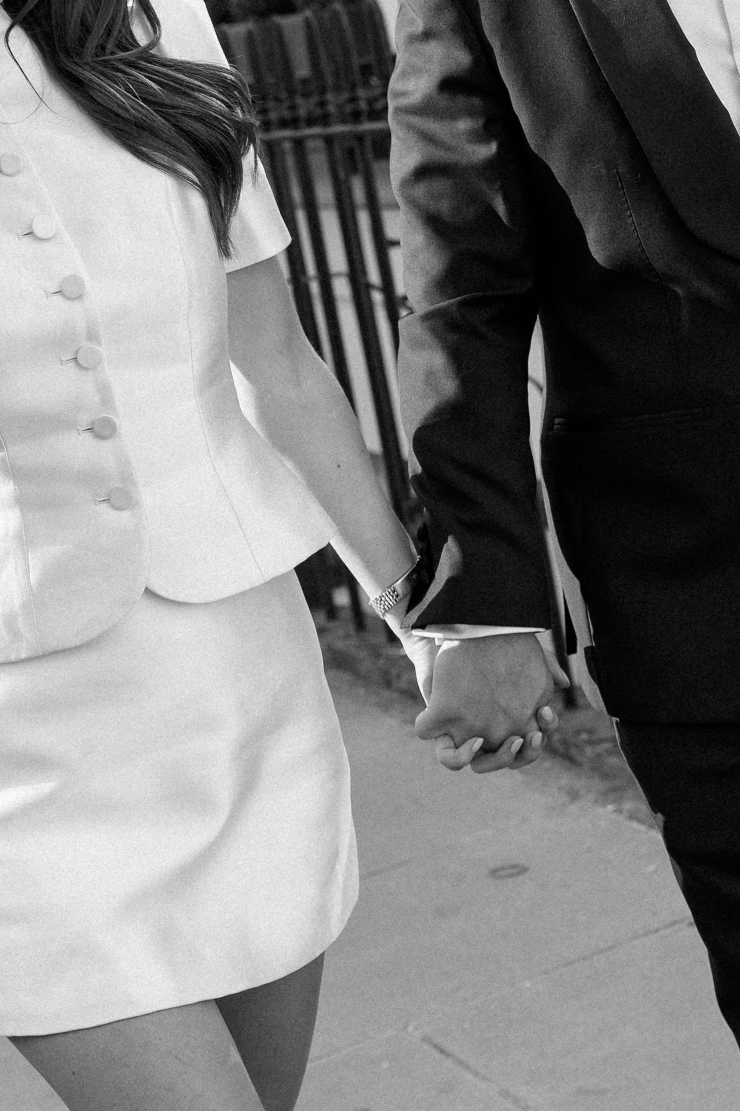 Black and white close-up of newlyweds holding hands, bride in white button-detail mini dress.