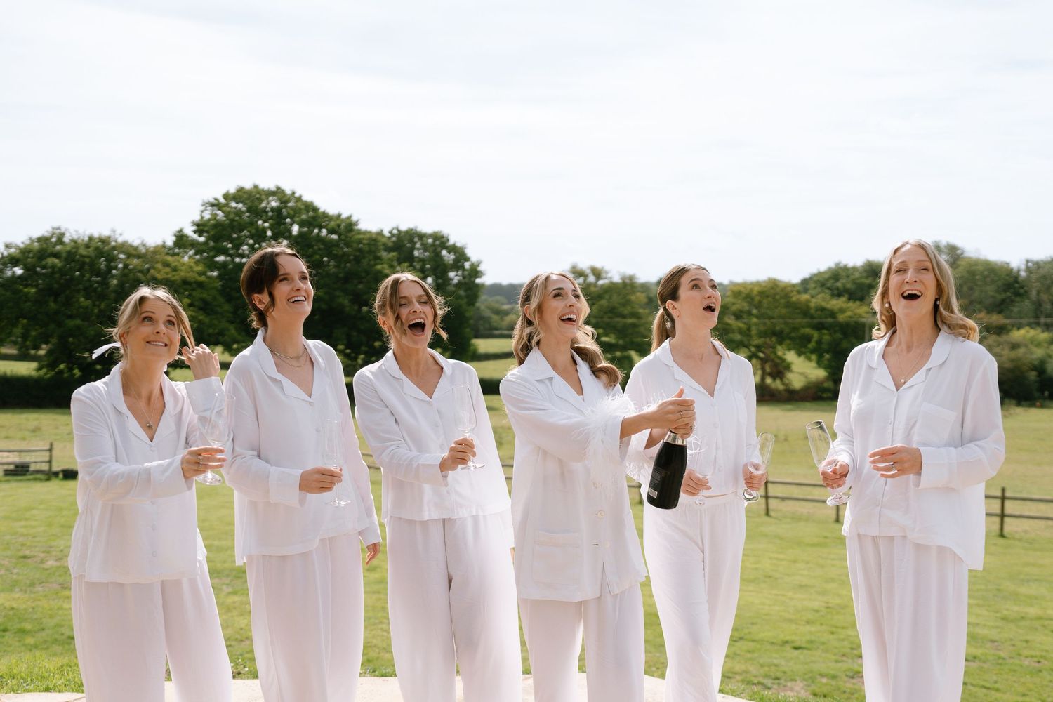 Six people in matching white outfits laugh together during an outdoor celebration.