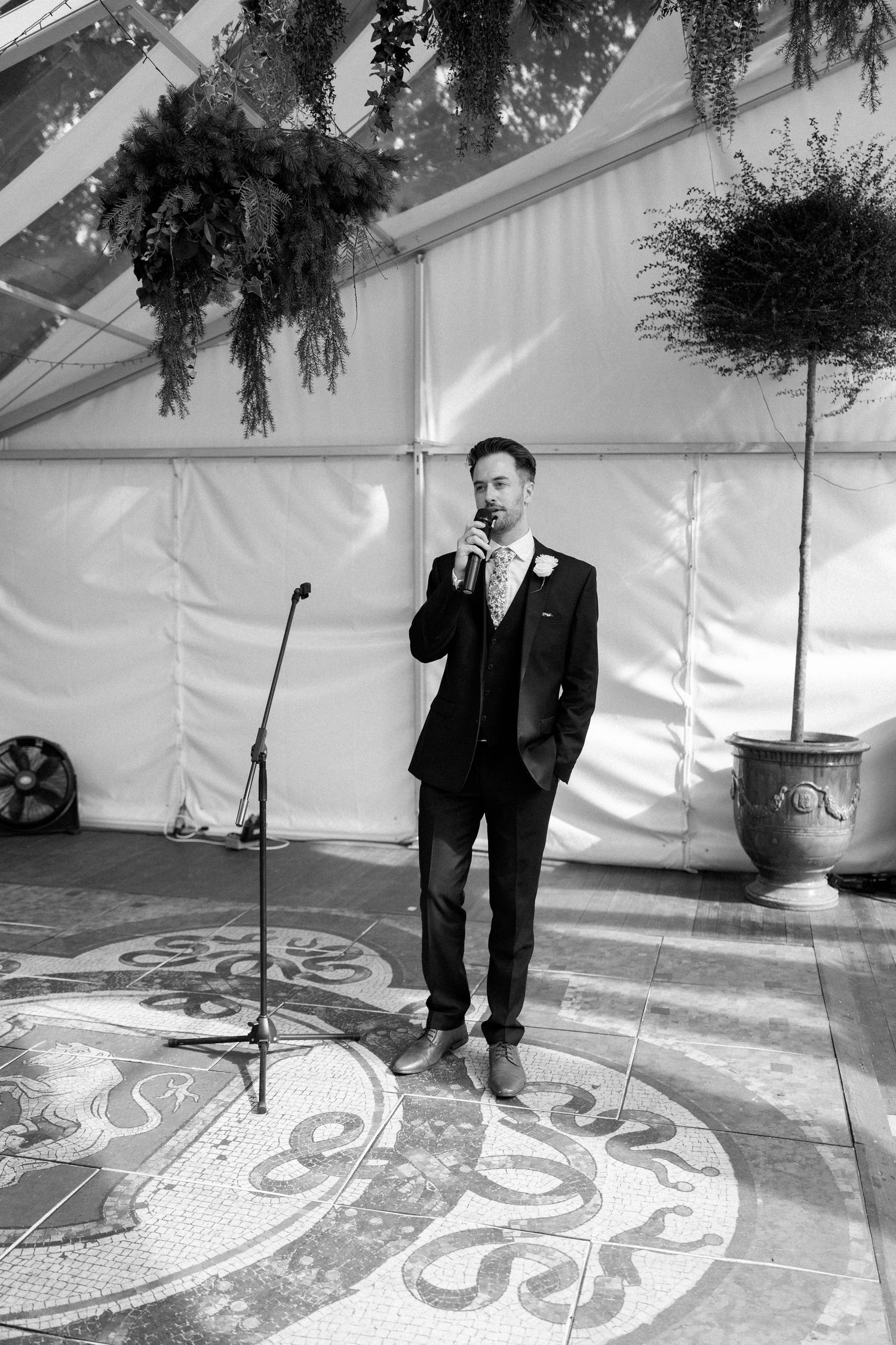 A speaker delivers remarks at a wedding reception on a decorated floor under a white tent.