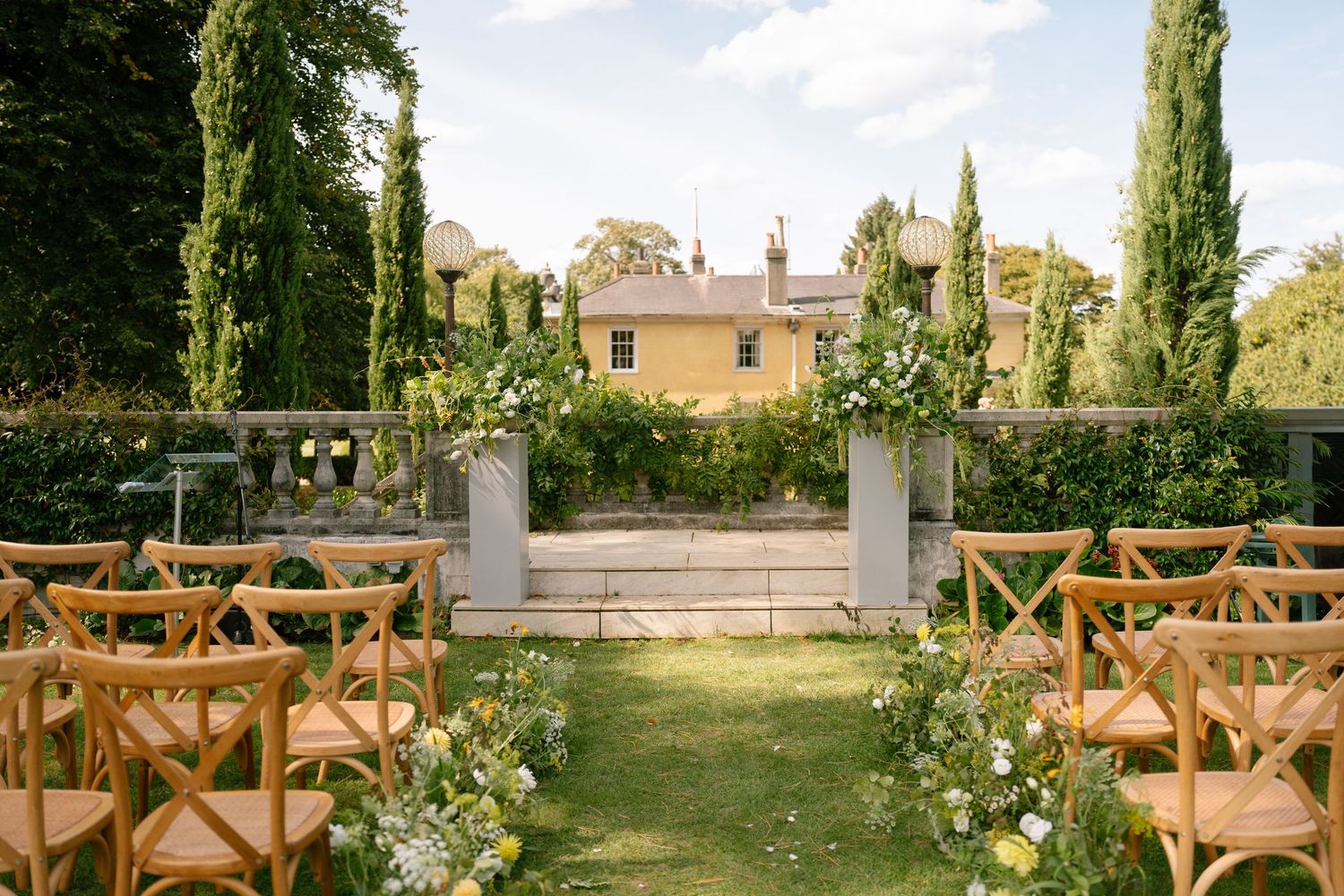 Outdoor wedding ceremony setup with wooden cross-back chairs and floral decorations.