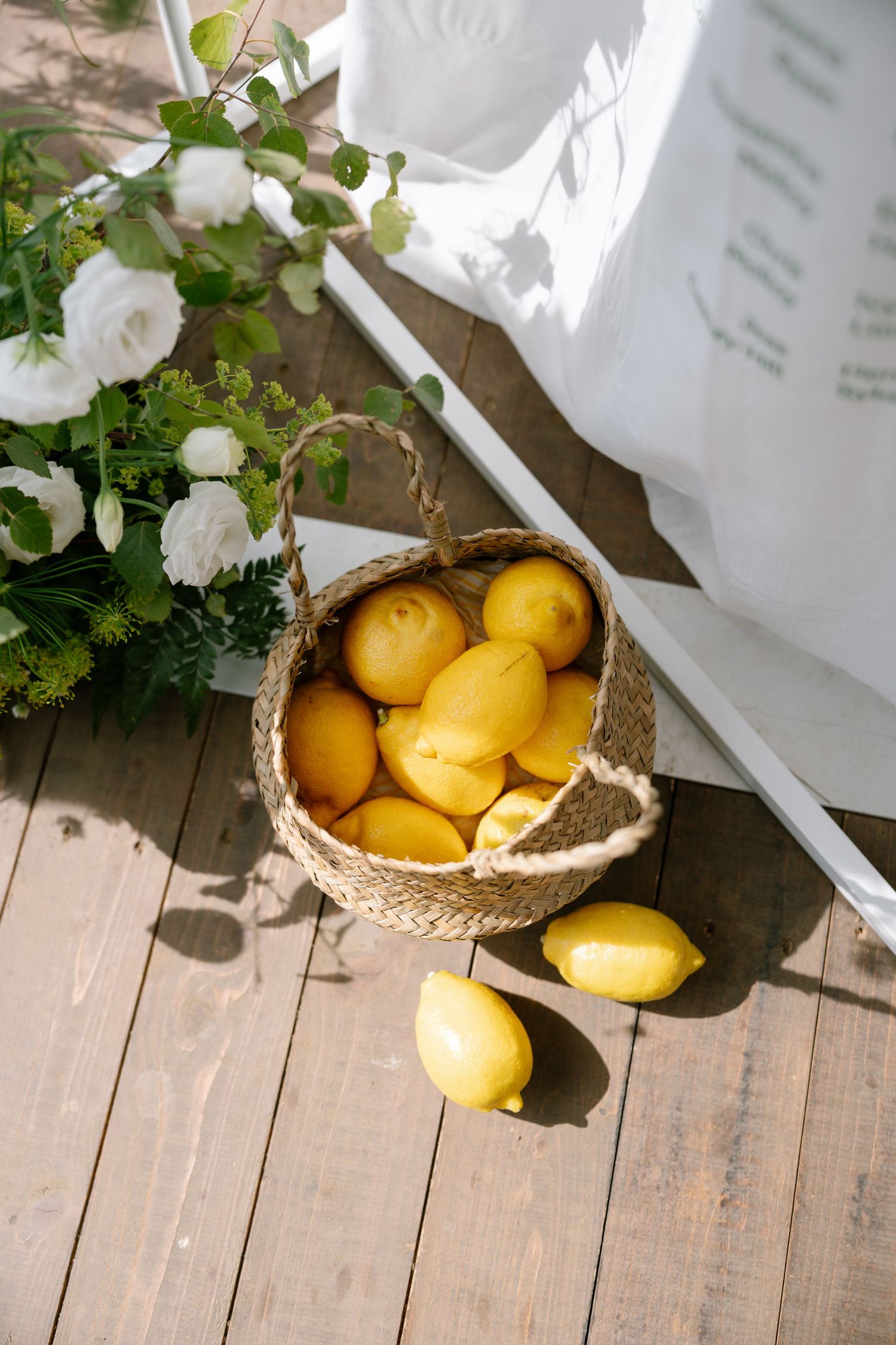 Fresh yellow lemons displayed in a woven basket on wooden decking under white fabric.