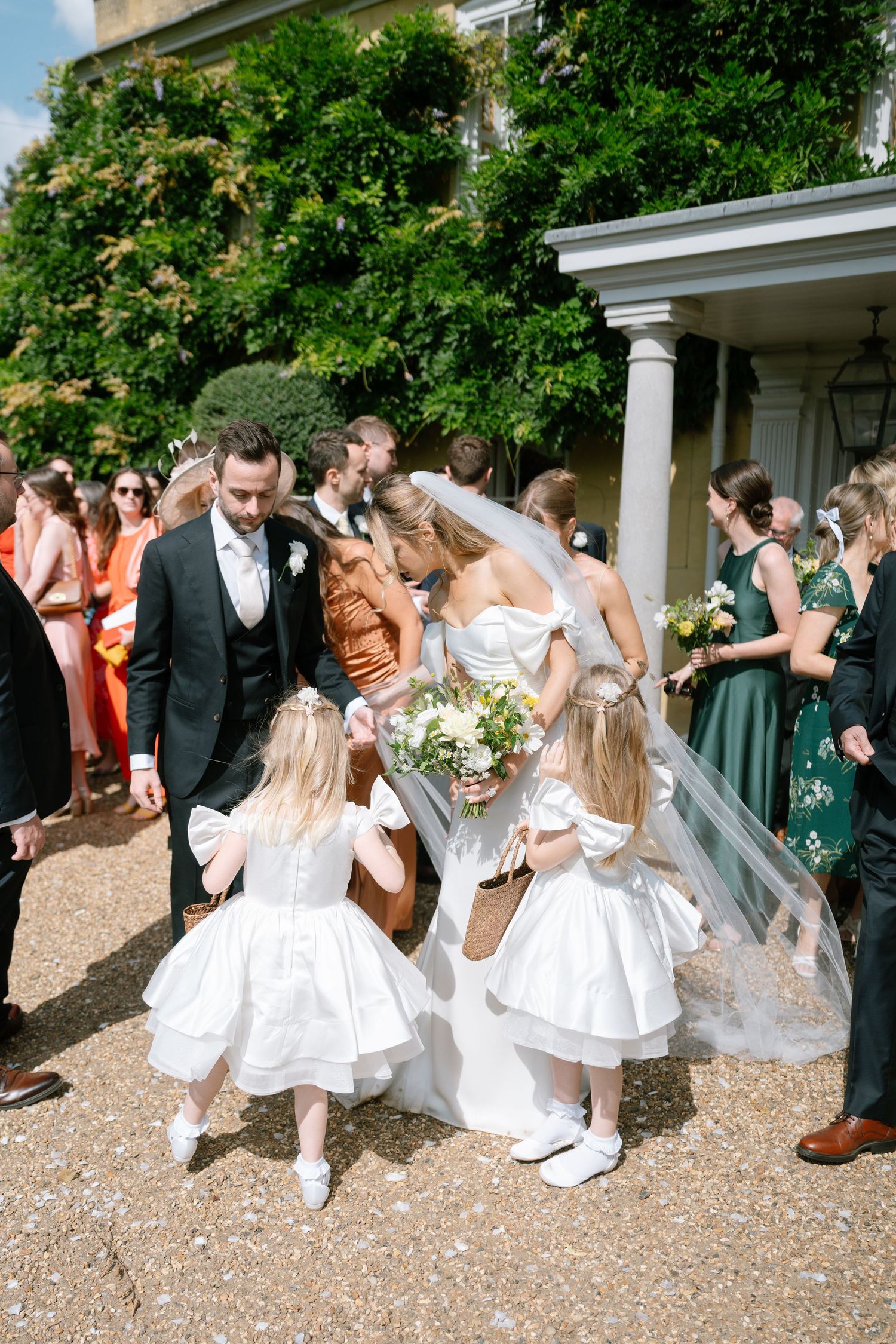 Wedding party celebrates on gravel path with guests throwing confetti outside venue.